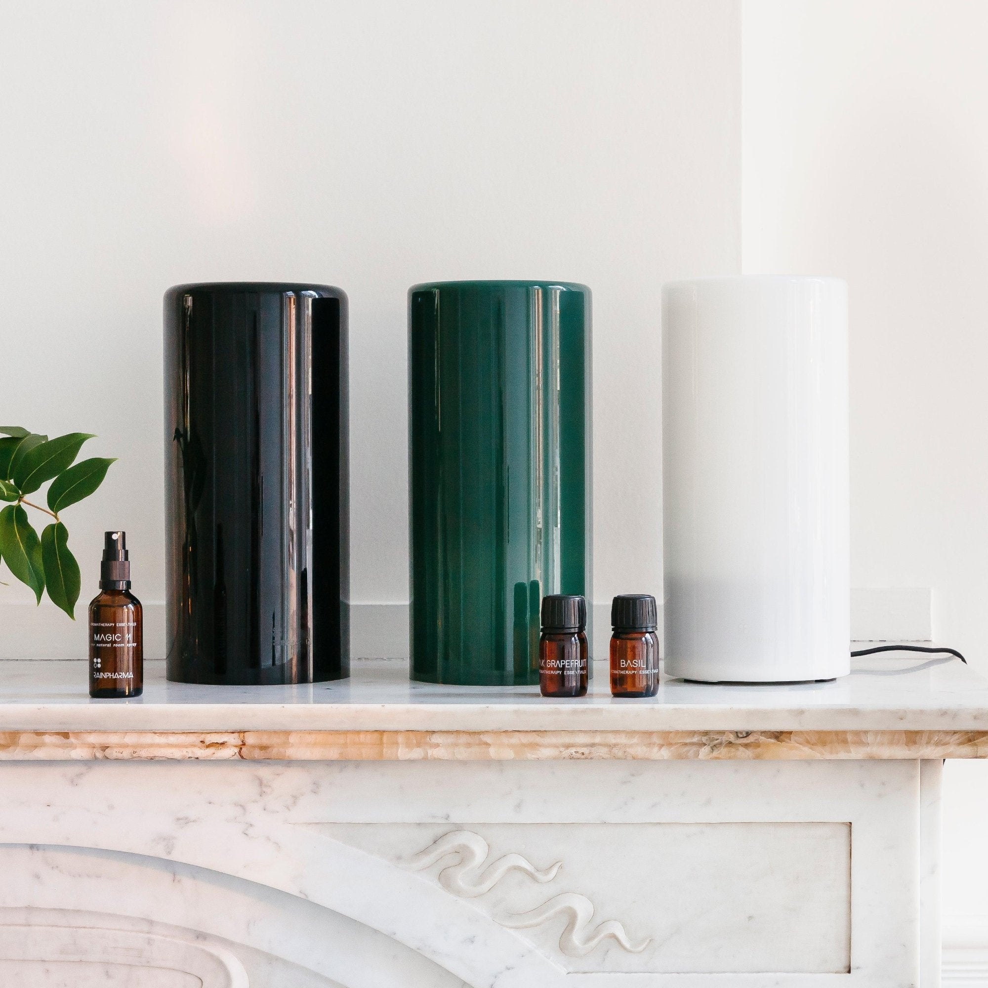 De Aroma Diffuser - Aurora Glass Sleeve van RainPharma, verkrijgbaar in zwart, groen en wit, staat sierlijk op marmer naast vier bruine flessen met essentiële oliën. Het voegt sfeervol licht toe voor een verbeterde geurverspreiding en ambiance in elke kamer.
