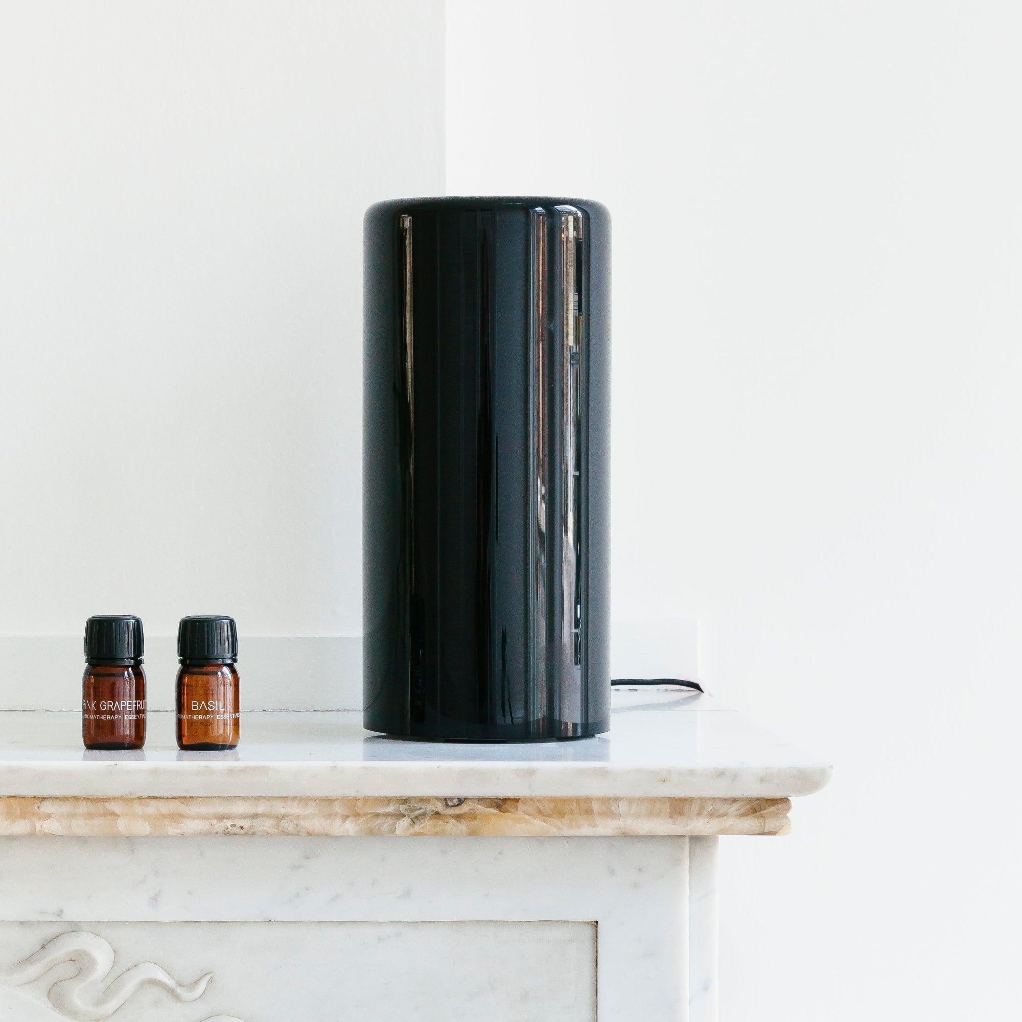 De RainPharma Aroma Diffuser - Aurora Glass Sleeve (in strak zwart) staat op een wit marmeren oppervlak naast twee kleine bruine flesjes met de labels "BASIL" en "X GROVE OIL" en biedt een sfeervolle, lichte en moeiteloze geurverspreiding.