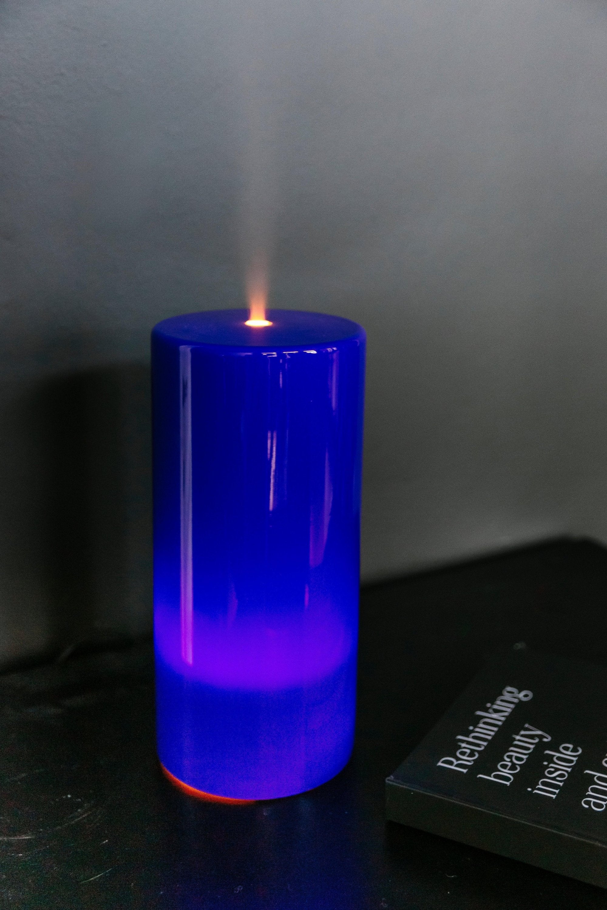 Een hoge, glimmende blauwe kaars verspreidt een kleine vlam op een donker oppervlak naast een gedeeltelijk zichtbaar boek met tekst op de omslag. Hierdoor ontstaat een sfeer die vergelijkbaar is met die van de RainPharma Aroma Diffuser - Aurora Glass Sleeve.