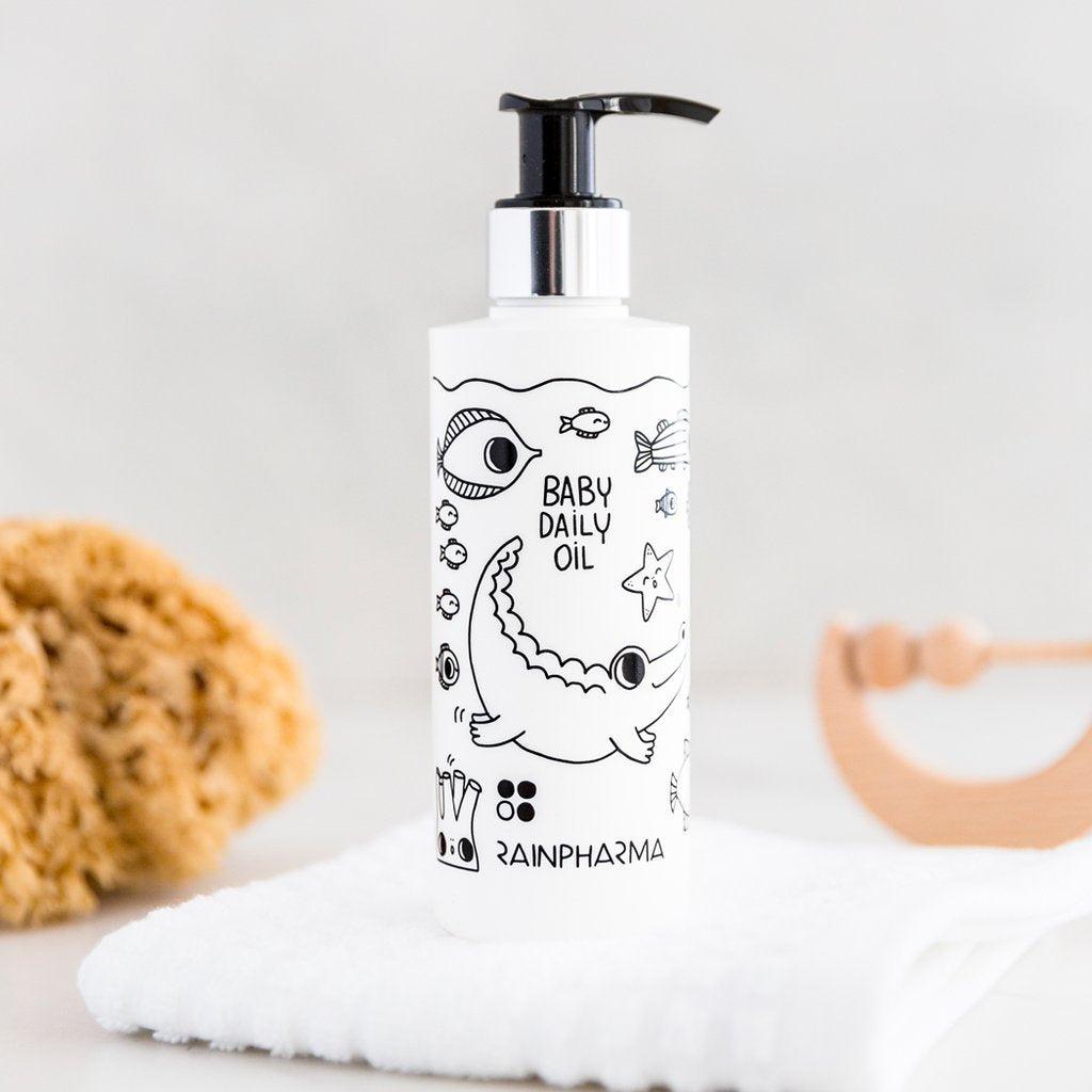 Een 200 ml fles RainPharma Baby Daily Oil voor hypoallergene verzorging rust op een opgevouwen witte handdoek, met een spons en houten babyspeeltje in de buurt.