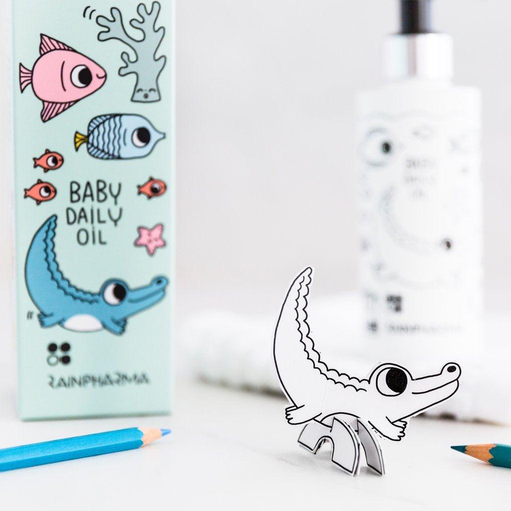 Een flesje en doosje RainPharma Baby Daily Oil - 200 ml, met kleurrijke cartooneske dieren zoals een krokodil en een kleine uitgeknipte krokodil op een wit oppervlak met kleurpotloden.
