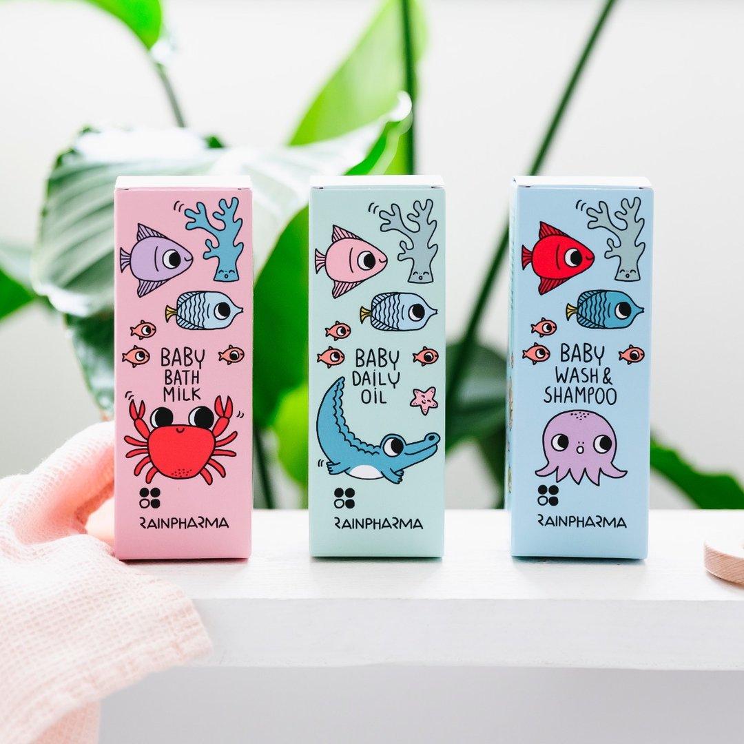 Drie dozen met RainPharma-babyproducten - Badmelk, Baby Dagelijkse Olie (200 ml) en Was & Shampoo - elk met cartoonachtige zeedieren op pastelkleurige verpakkingen, uitgestald op een wit oppervlak.