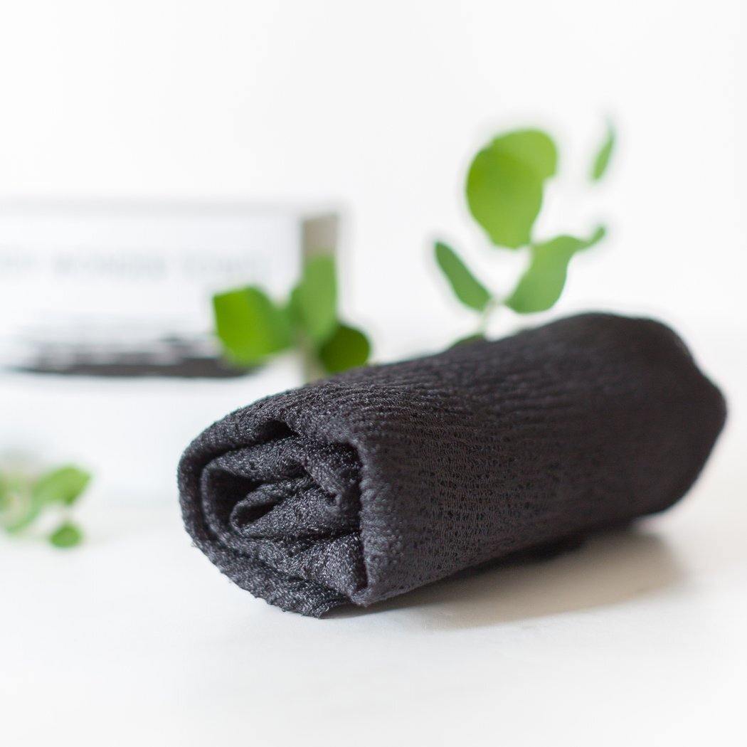 Een opgerolde, zwarte, gestructureerde doek die lijkt op een Body Wonder Towel van RainPharma is te zien, met wazig groen en een verpakking plantaardige douchegel op de achtergrond.