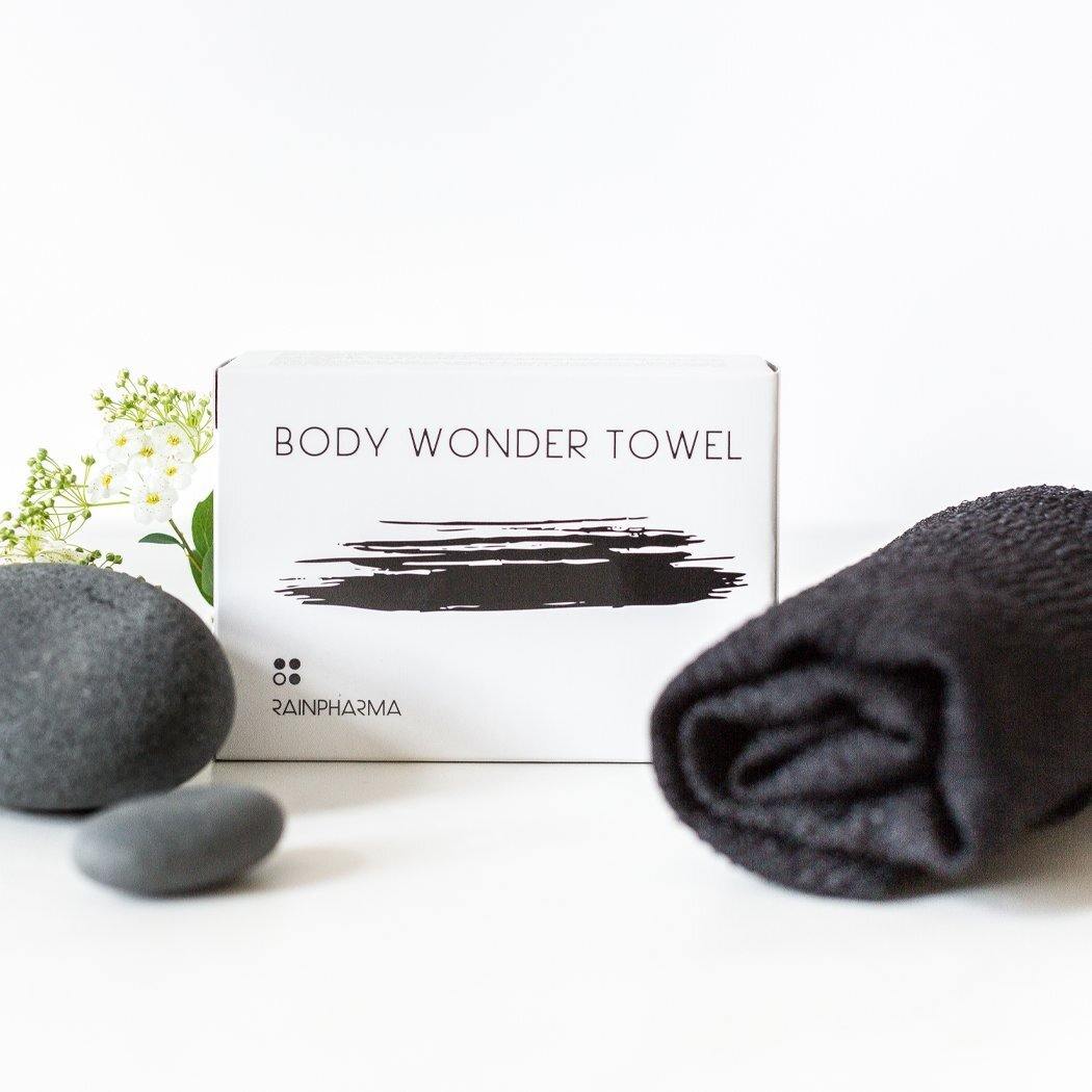 Naast een doosje met het opschrift RainPharma Body Wonder Towel liggen een exfoliërende wasdoek, een opgerolde zwarte handdoek, gladde steentjes en delicate witte bloemen op een maagdelijk witte achtergrond.