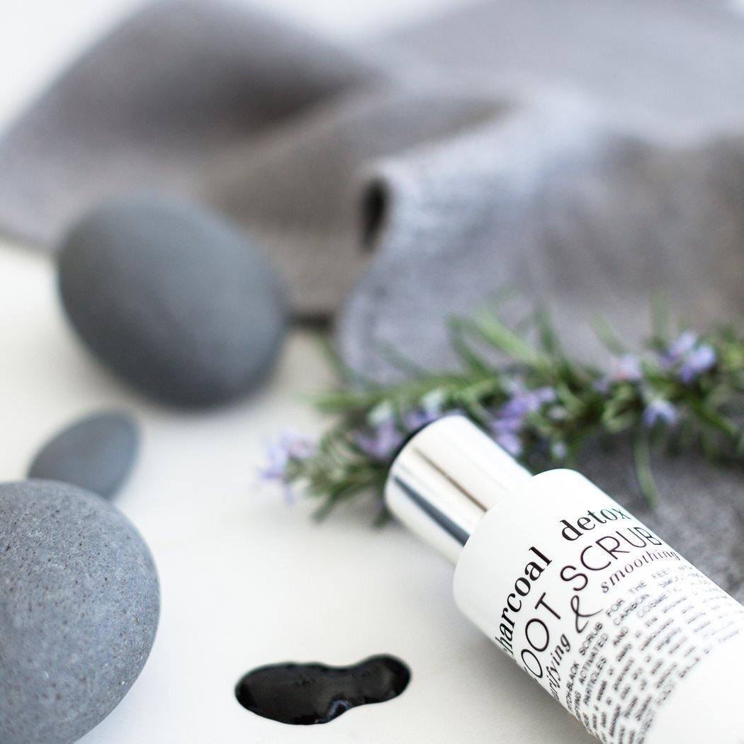 Een fles RainPharma Charcoal Detox Foot Scrub (100 ml) staat naast gladde grijze stenen, een grijze doek, verse kruiden en een klein plasje zwarte vloeistof.
