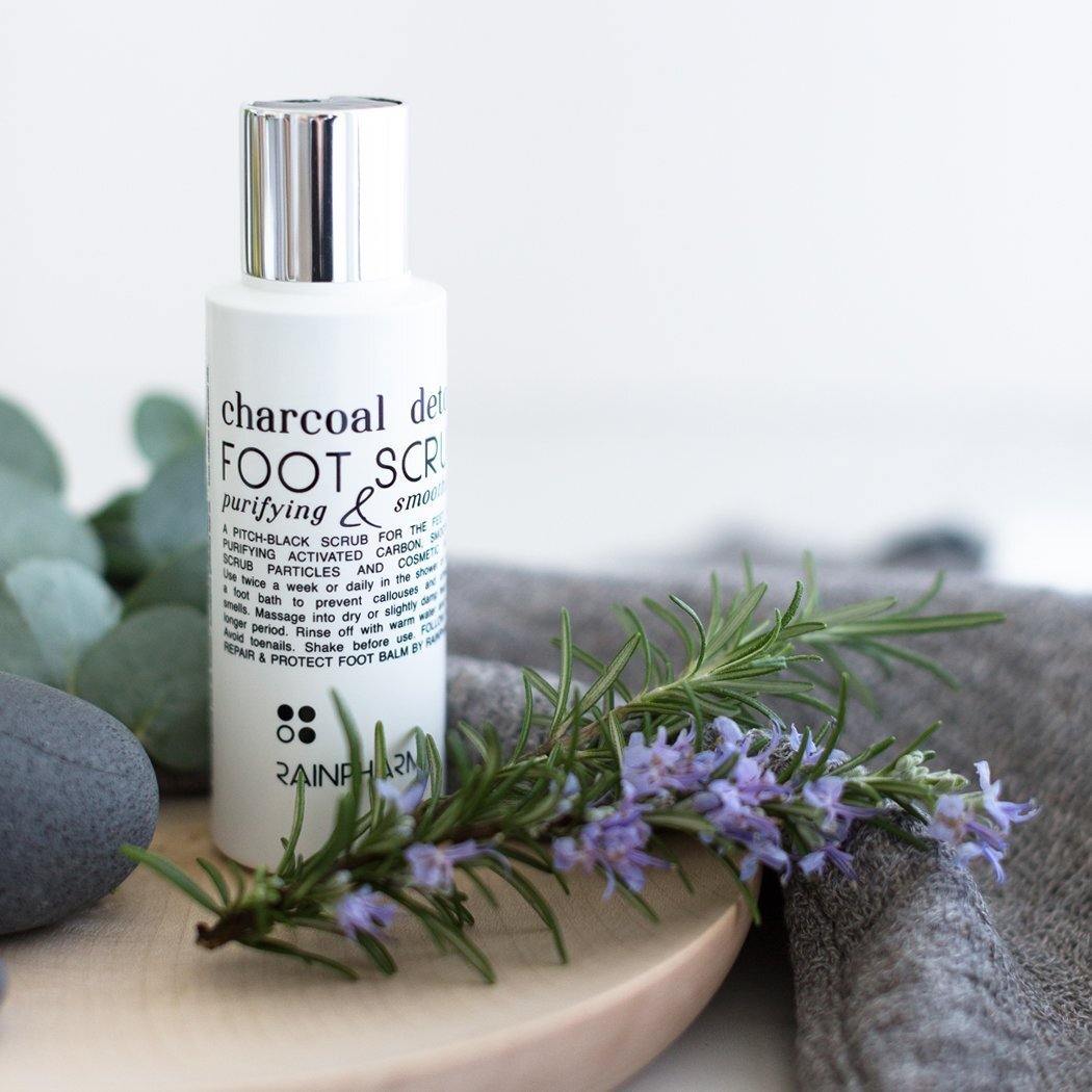 Een fles RainPharma Charcoal Detox Voetscrub (100 ml) staat op een licht oppervlak naast takjes rozemarijn, een grijze handdoek en gladde stenen.