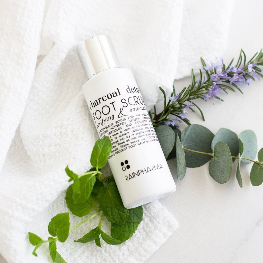 Een fles RainPharma Charcoal Detox Foot Scrub (100 ml) staat op een witte handdoek naast verse munt-, eucalyptus- en rozemarijntakjes.