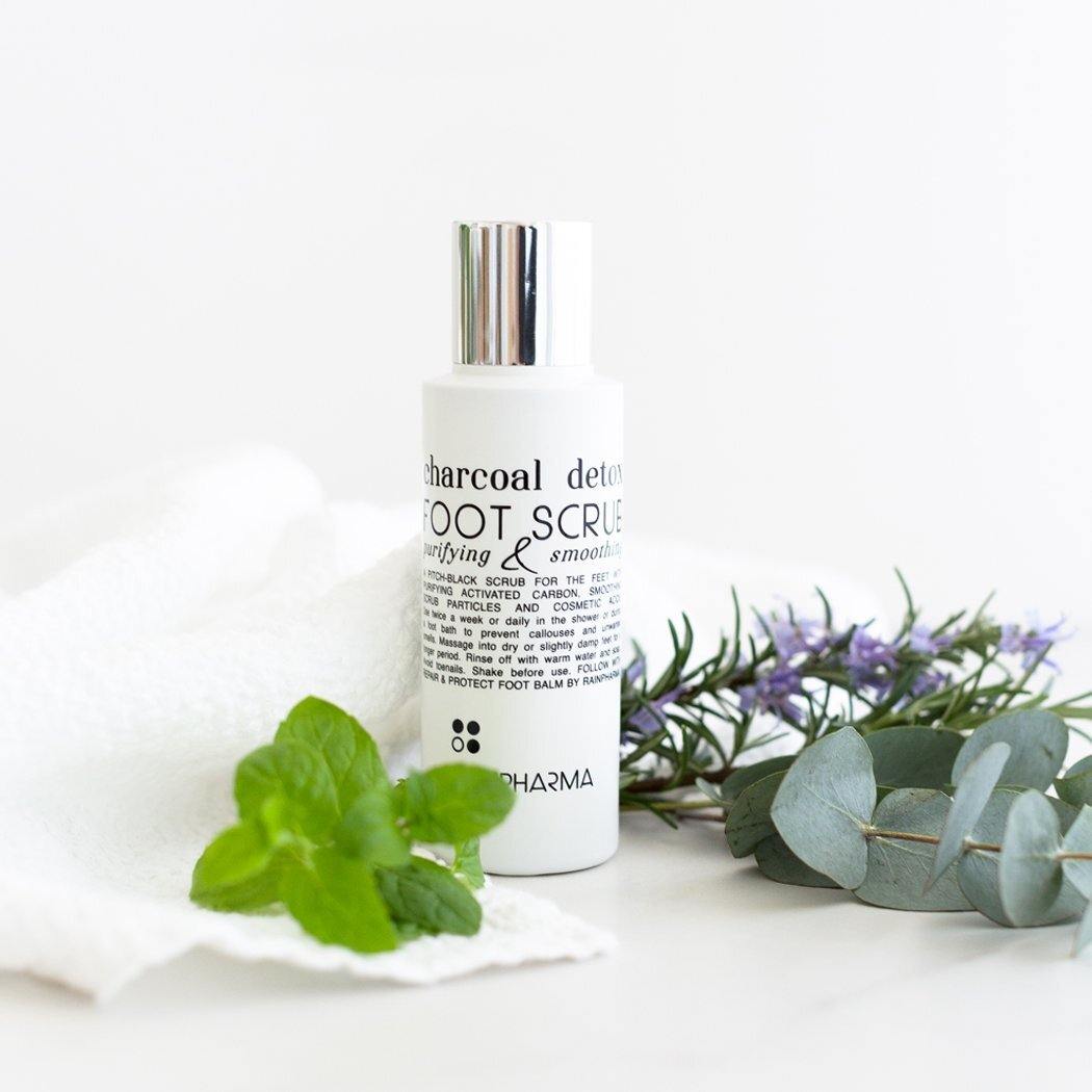 Een fles RainPharma Charcoal Detox Foot Scrub (100 ml) staat op een wit oppervlak naast een witte handdoek, verse muntblaadjes en takjes rozemarijn en eucalyptus.