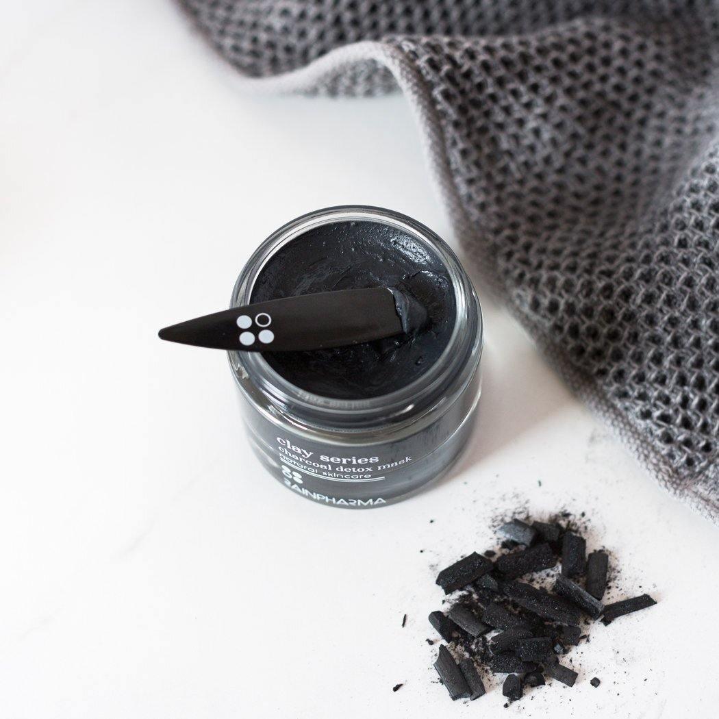 Een geopend RainPharma Charcoal (Detox) Mask - Zuiverend masker elk huidtype potje met een zwarte applicator staat op een wit oppervlak in de buurt van stukjes houtskool en een grijze handdoek - perfect voor een vette huid die zuivering zoekt.