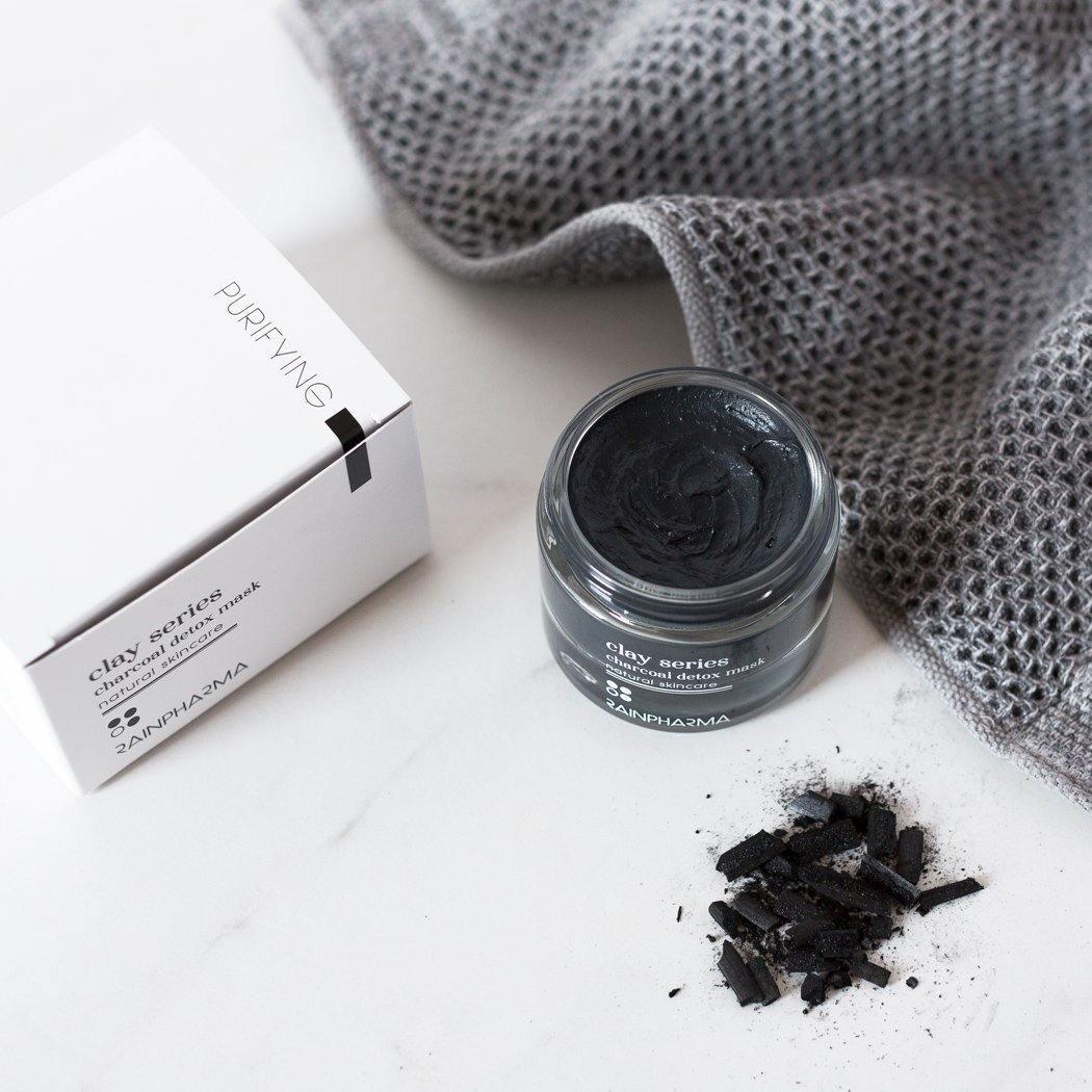 Een pot RainPharma Charcoal (Detox) Mask - het zuiverende masker voor elk huidtype - staat naast de doos, met stukjes houtskool en een grijze handdoek op een witte ondergrond.
