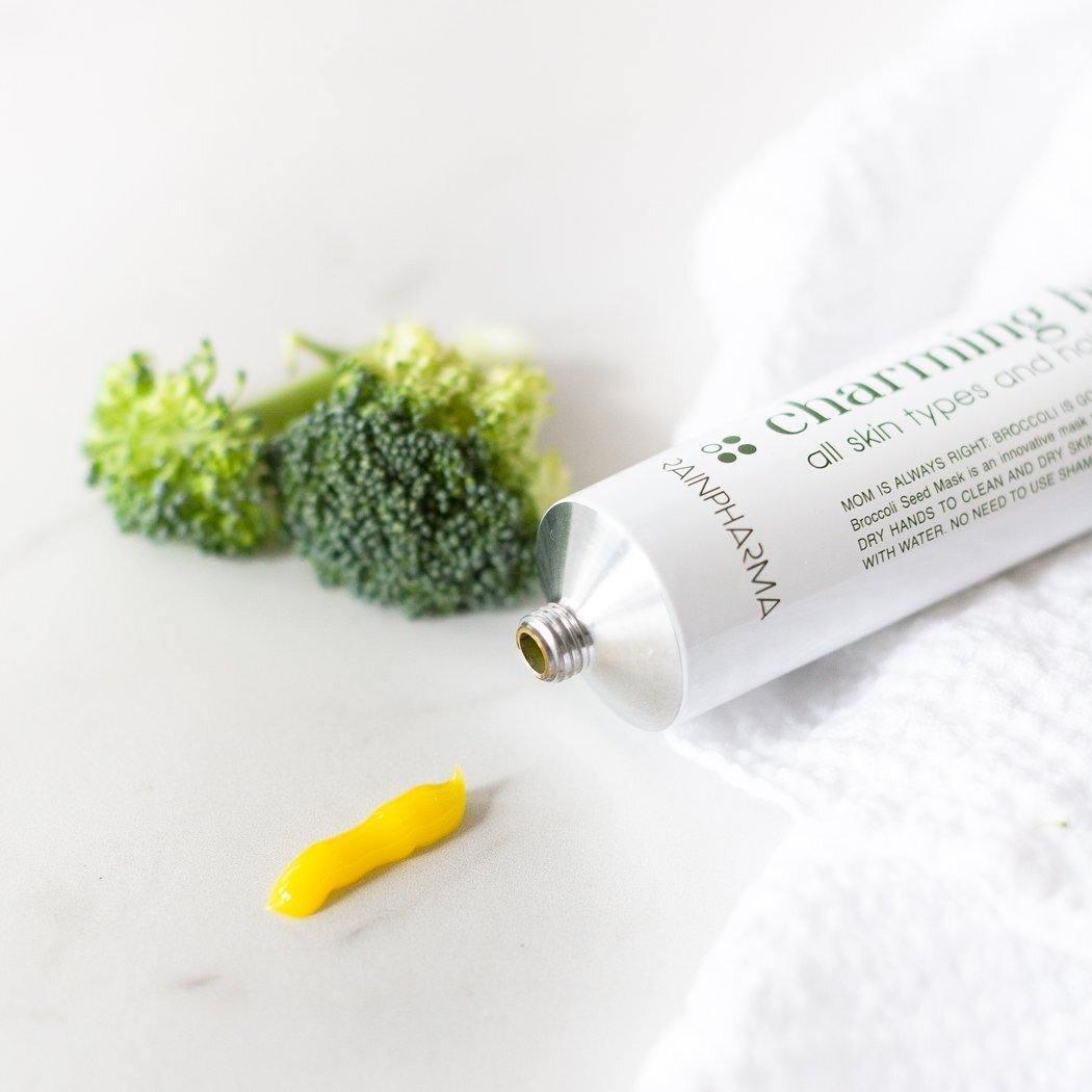 Een geopende tube RainPharma Charming Broccoli Seed Mask - kalmerend en herstellend, met gele crème, zit naast broccoliroosjes op een wit oppervlak, wat de superfood huidverzorging vibe benadrukt; een handdoek is gedeeltelijk zichtbaar.