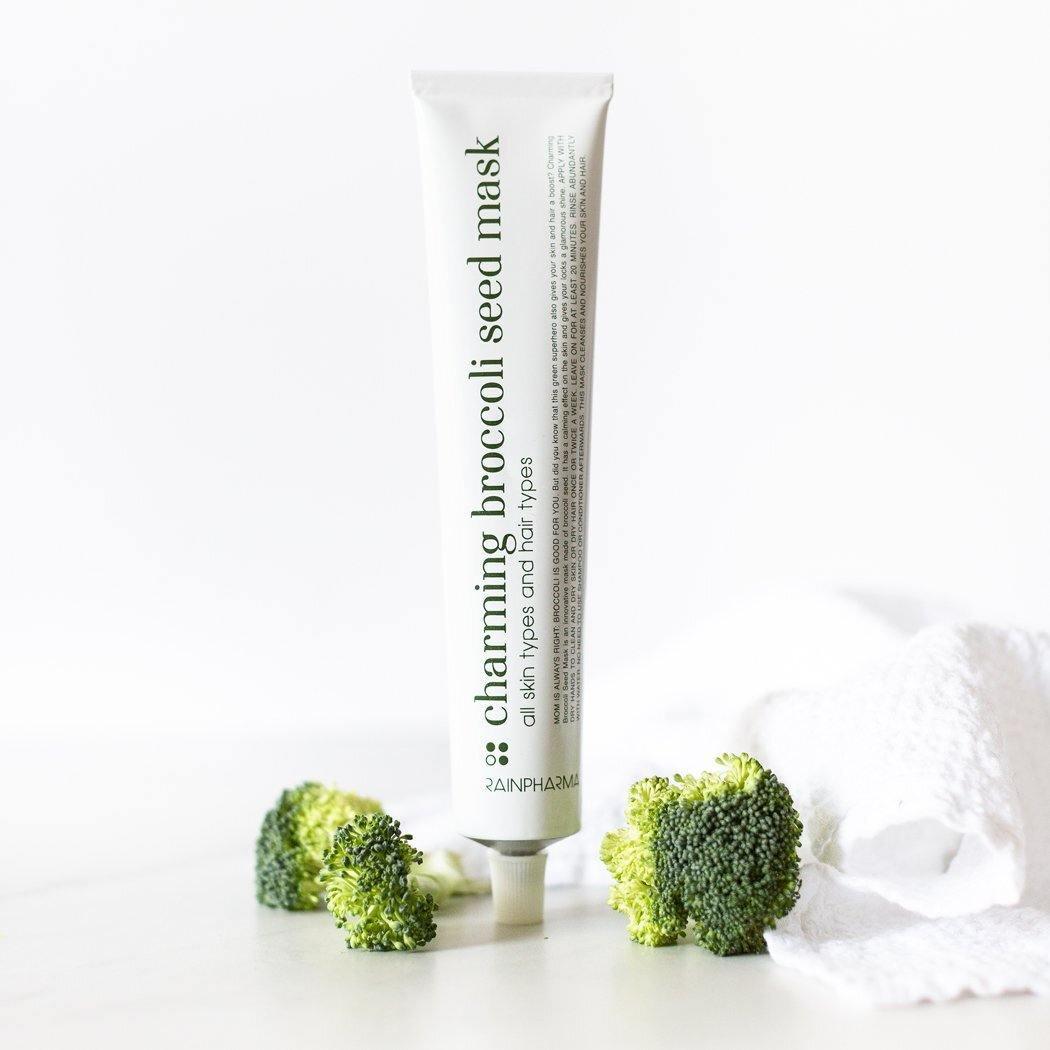 Een tube Charming Broccoli Seed Mask van RainPharma - kalmerend en herstellend staat rechtop op een wit oppervlak met broccoli en een handdoek, perfect als een broccolizaad masker voor superfood huidverzorging.