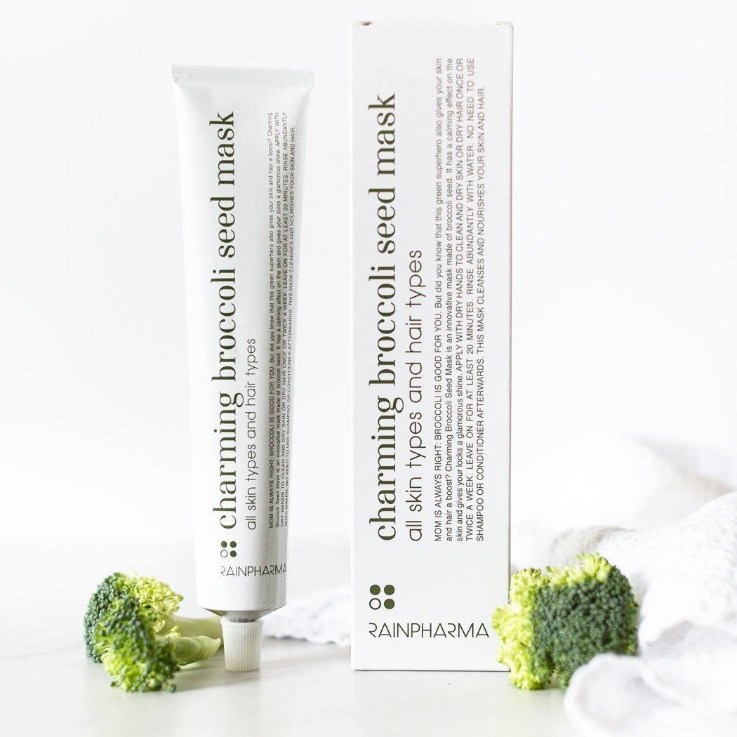 Een tube en doosje RainPharma Charming Broccoli Seed Mask - kalmerend en herstellend staan rechtop op een wit vlak, met twee broccoliroosjes ernaast.