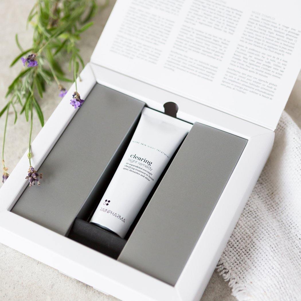 Een witte tube RainPharma Clearing Night Remedy dagcrème voor onzuivere of vette huid wordt getoond in een open wit doosje met schuimrubberen inzetstukken, naast lavendeltakjes en een wit doekje met structuur.