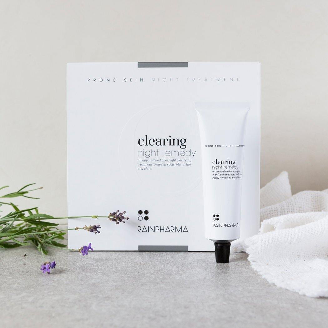 Een witte tube van RainPharma Clearing Night Remedy - Dagcrème voor onzuivere of vette huid staat voor zijn doosje, met lavendeltakjes en een getextureerd wit doekje op een licht oppervlak - perfect voor de vette huid en rijk aan natuurlijke ingrediënten.
