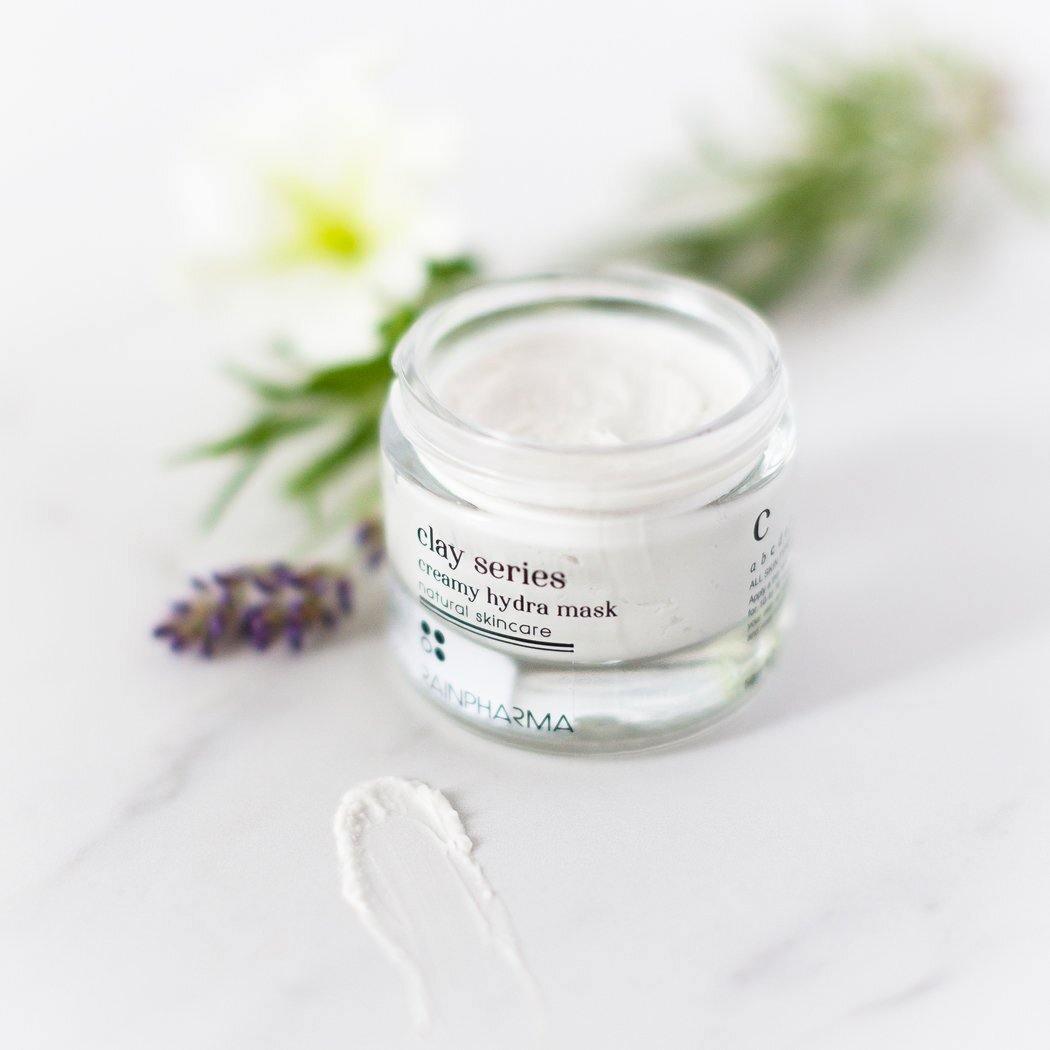 Een potje Creamy Hydra Mask - hydratatie van RainPharma ligt op een oppervlak met een klein beetje crème op de voorgrond en wazige bloemen op de achtergrond, waardoor de natuurlijke oorsprong wordt benadrukt.