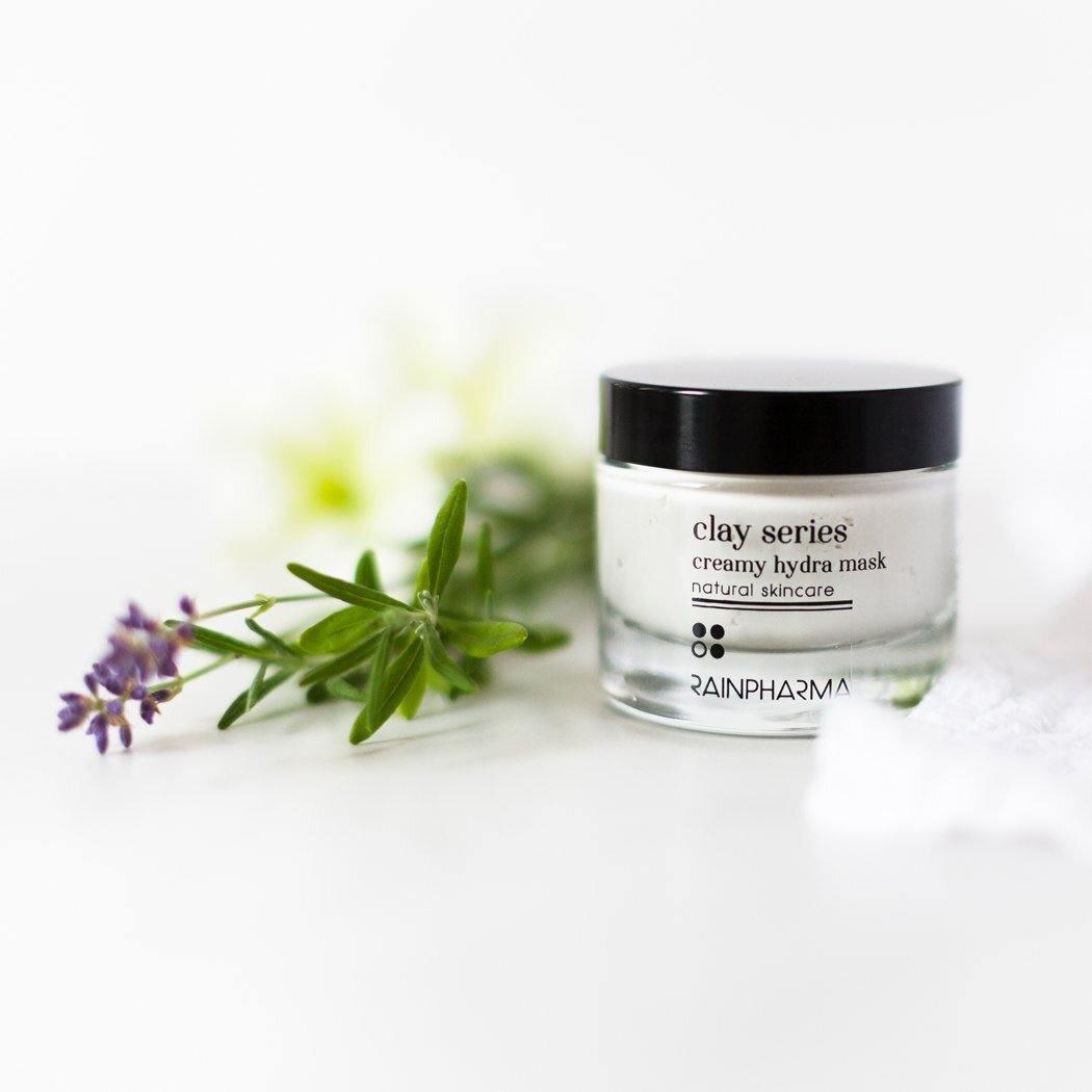 Een potje Creamy Hydra Mask - hydratatie van RainPharma, geroemd om zijn plantaardige oorsprong, staat op een wit oppervlak versierd met groene takjes en kleine paarse bloemetjes.