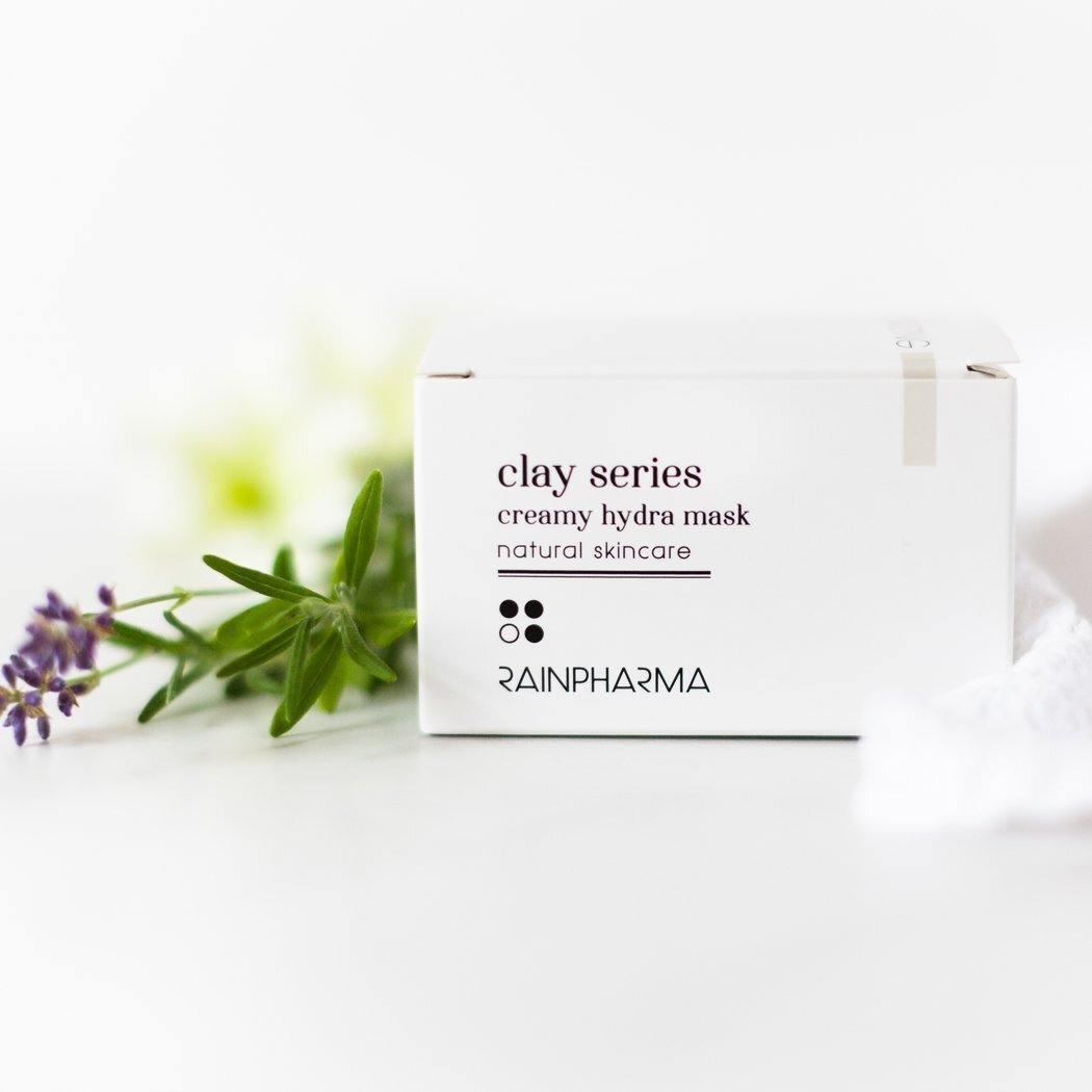 Een RainPharma Creamy Hydra Mask - hydratatie box staat op een wit oppervlak, vergezeld door lavendel en een groen takje. Het hydraterende kleimasker, gemaakt met plantaardige ingrediënten, wordt weergegeven tegen een helderwitte achtergrond.