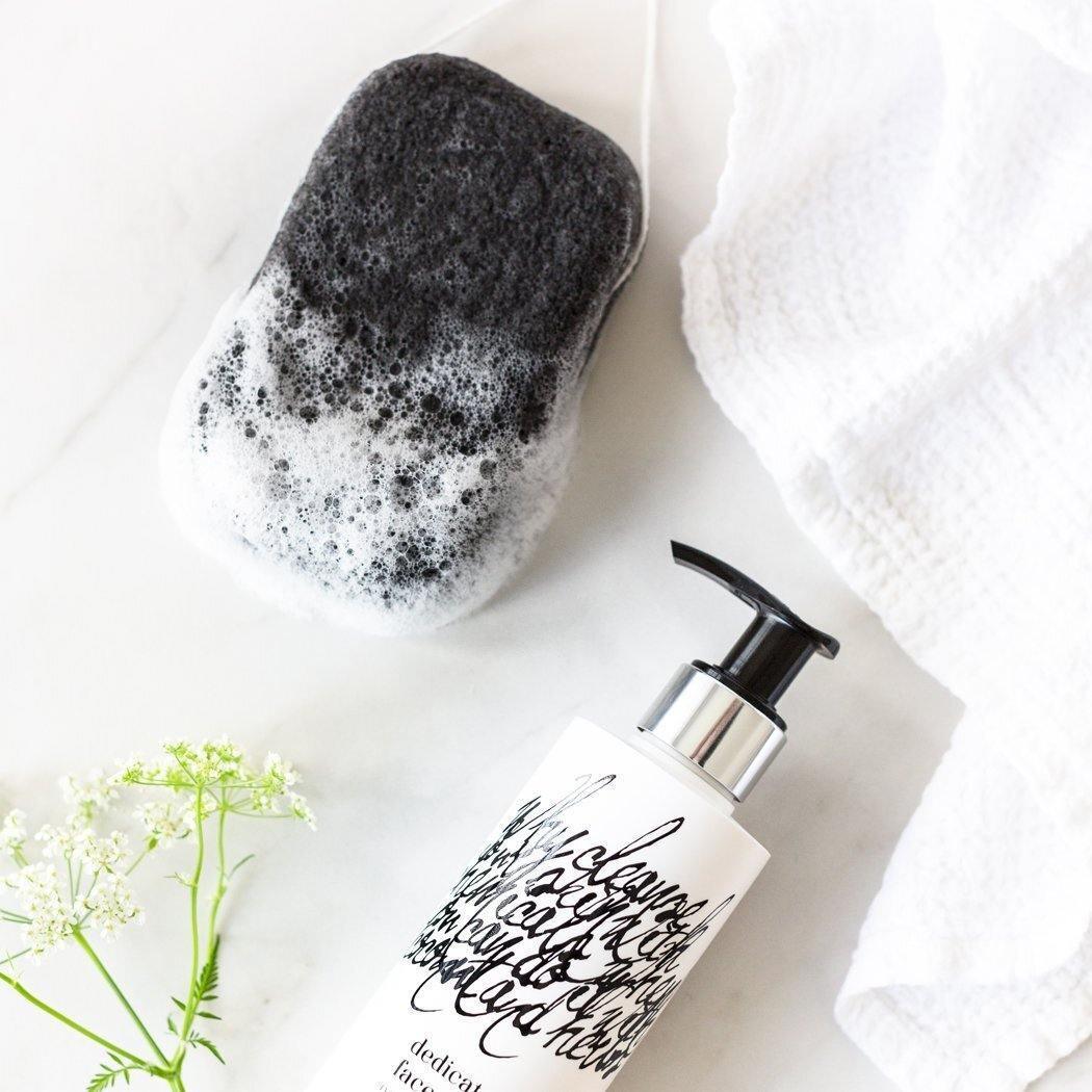 Een zwart-witte spons met zeepsop ligt naast een witte pompflacon RainPharma Dedicated Face Wash 200 ml en een witte handdoek op een marmeren oppervlak, met een klein wit bloemetje ernaast.