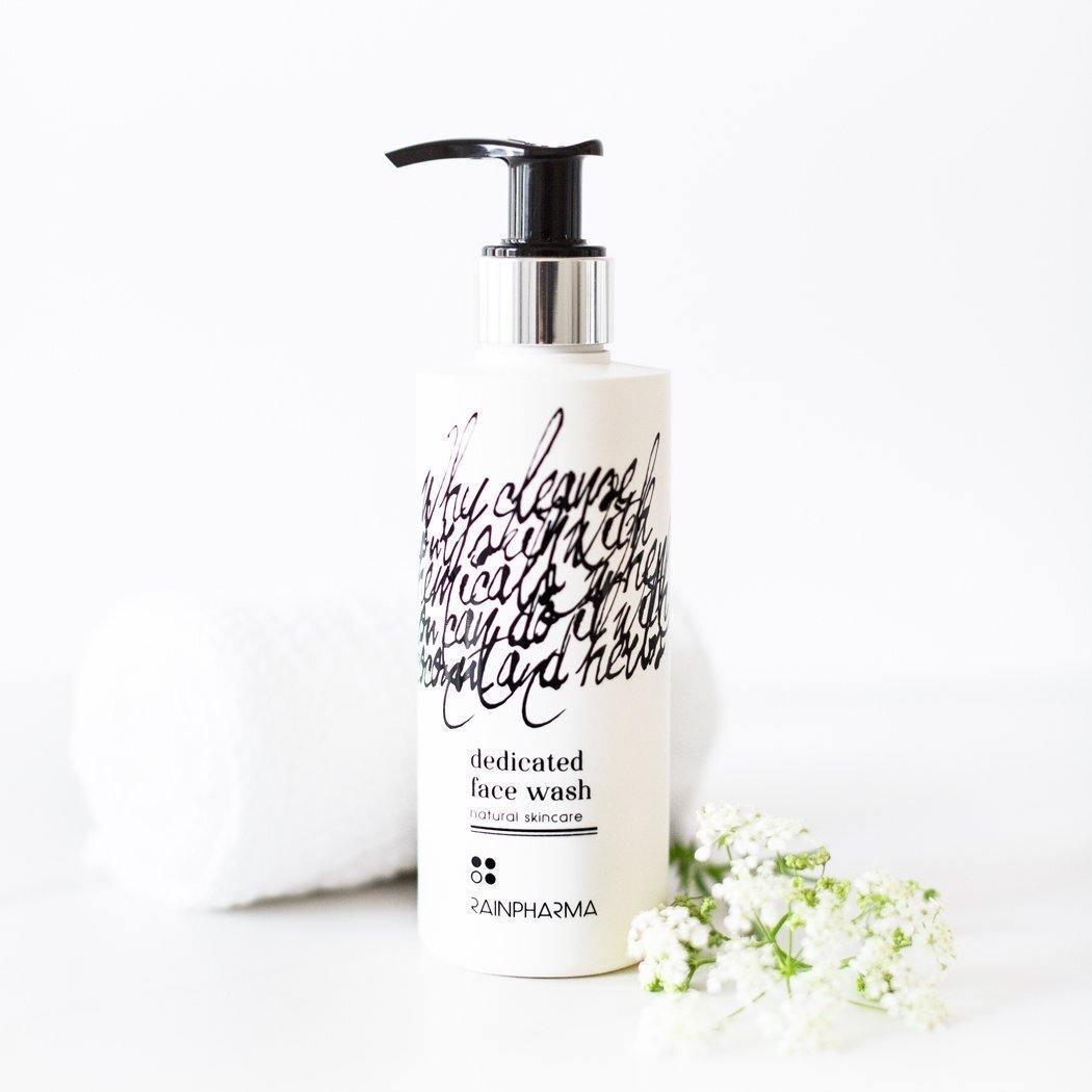 Een fles RainPharma Face Wash, een gezichtsreiniger op waterbasis met pompdispenser, staat afgebeeld op een wit oppervlak naast een opgerolde handdoek en witte bloemen.