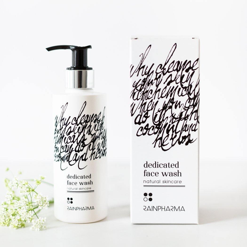 Een RainPharma Dedicated Face Wash-fles van 200 ml en een doos met abstract zwart opschrift op een witte verpakking worden afgebeeld naast een klein takje witte bloemen.