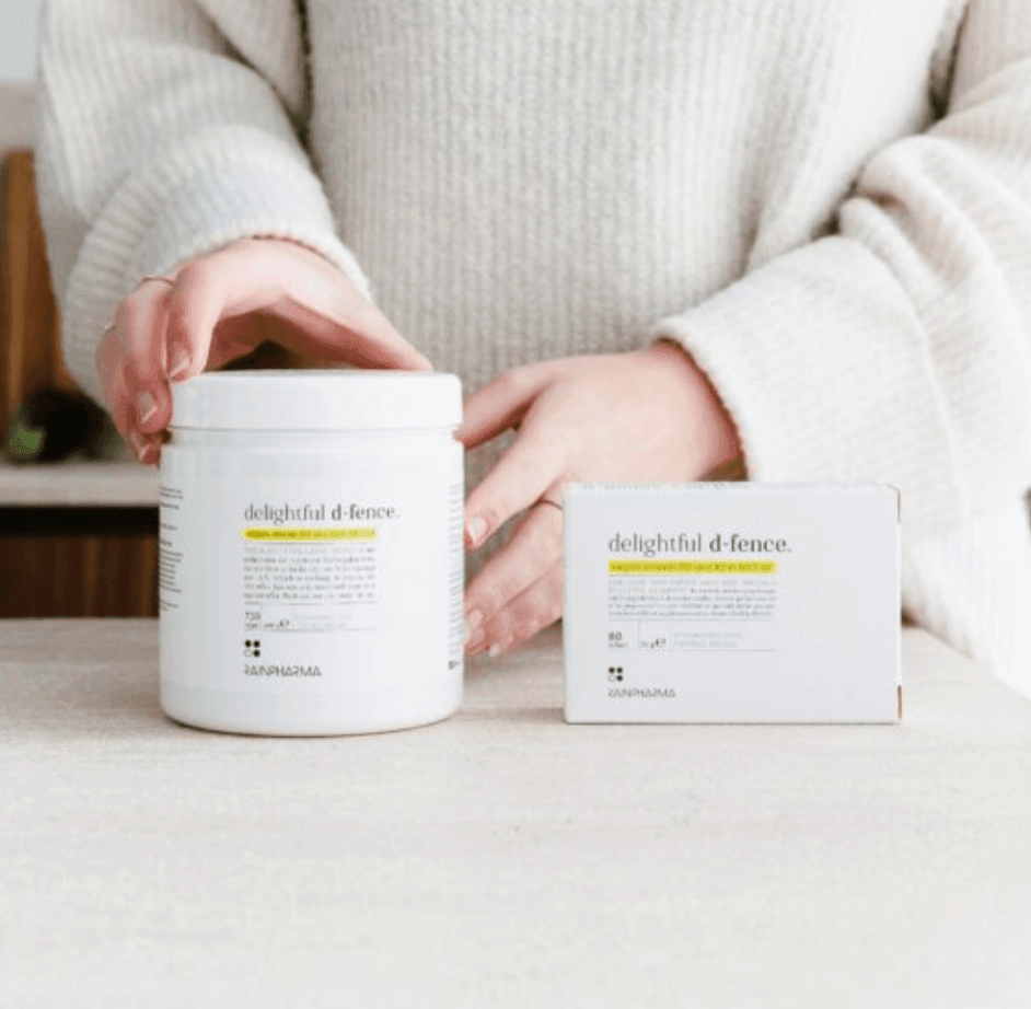 Een persoon houdt een witte pot met het opschrift "Delightful D-fence" uit de RainPharma Family Pack vast naast een bijpassend doosje op een aanrecht, wat het belang van vitamine D voor sterke botten onderstreept.