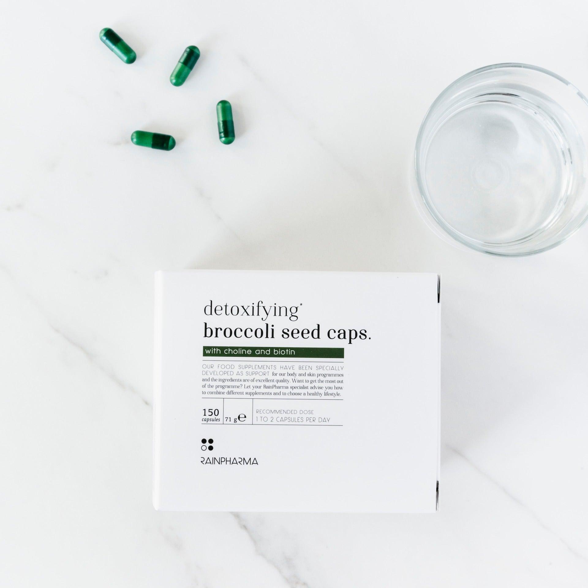 Een doosje RainPharma Detoxifying Broccoli Seed Caps, met broccolizaadextract en sulforafaan, staat op een wit oppervlak naast groene capsules en een glas water.