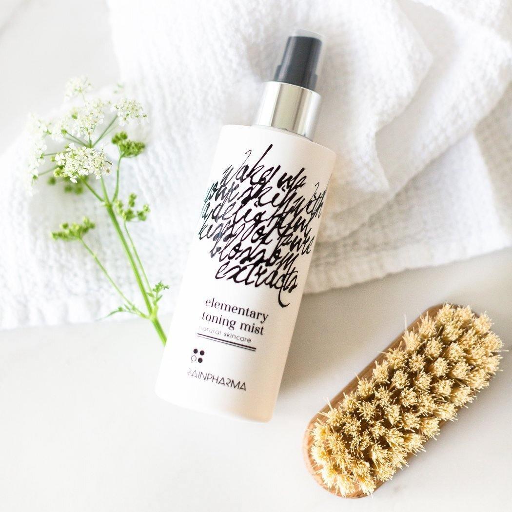 Een fles RainPharma Toning Mist staat op een wit oppervlak naast een lichaamsborstel, een zachte handdoek en een takje witte bloemen, waarmee deze favoriet in natuurlijke huidverzorging wordt benadrukt.
