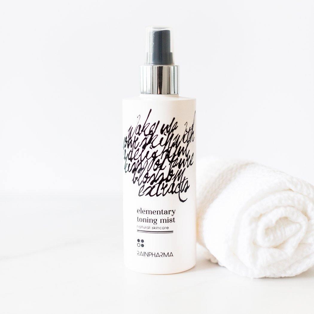 Een fles RainPharma Toning Mist (toner), een natuurlijke huidverzorging, staat naast een opgerolde witte handdoek op een wit oppervlak met een effen achtergrond.