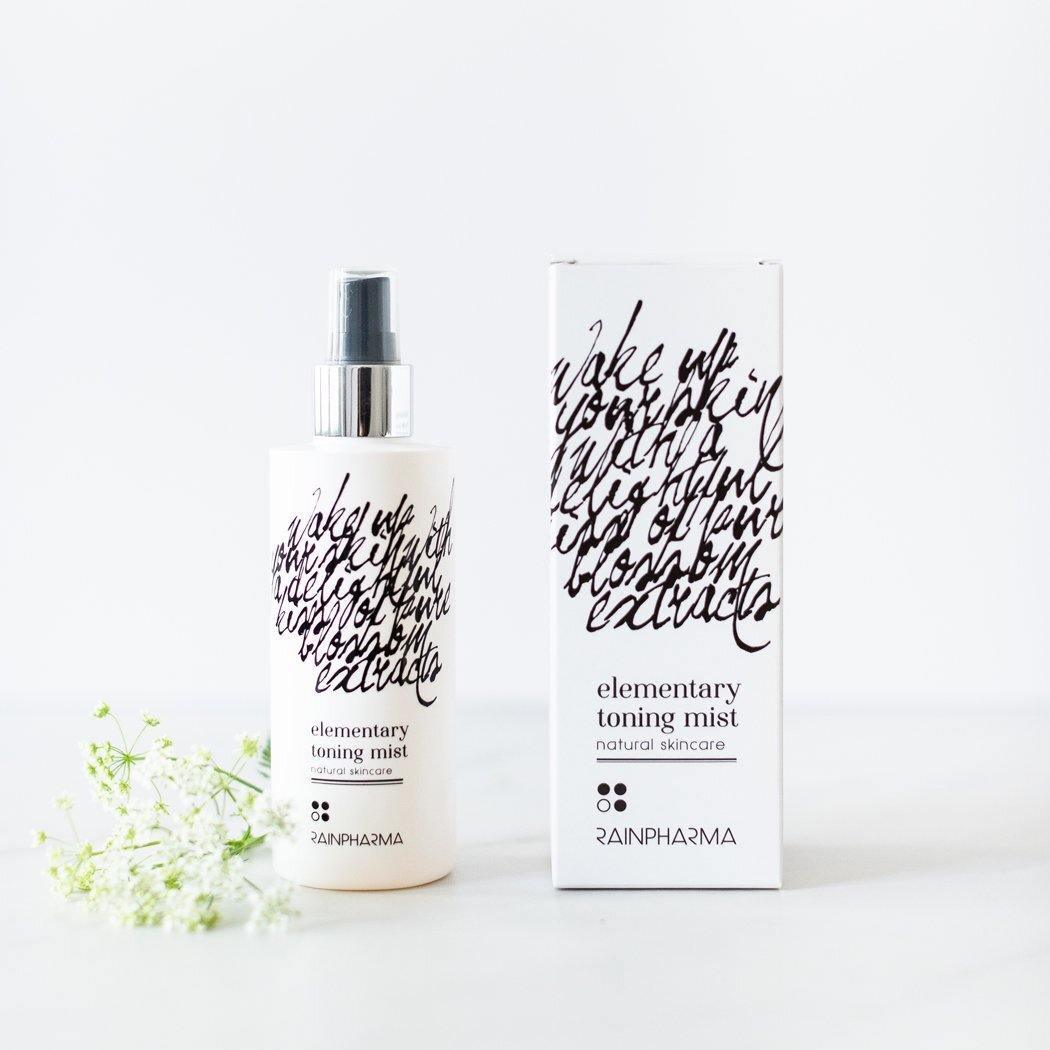Een flesje en doosje RainPharma Toning Mist toner staan naast elkaar op een wit oppervlak, vergezeld van een klein takje witte bloemen.
