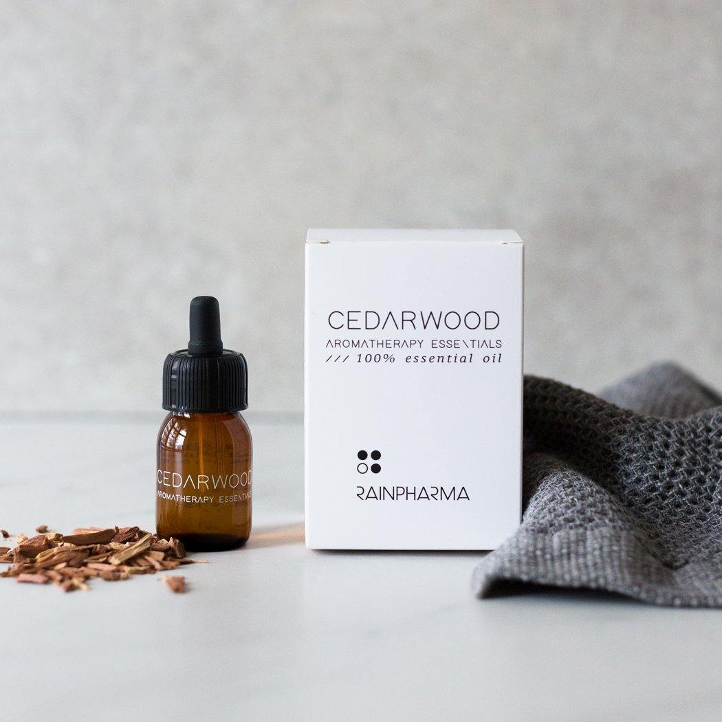 Een flesje RainPharma Essential Oil Cedarwood zit bij een wit doosje "Cedarwood Aromatherapy Essentials", een grijs doekje en cederhoutkrullen - perfect om een warme cedergeur in je aromadiffuser te brengen.