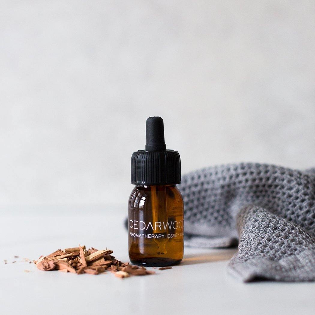 Een klein amberkleurig flesje RainPharma Essential Oil Cedarwood met een druppeldop rust naast cederhoutsnippers en een grijs doekje, ideaal om je aromadiffuser te vullen met de warme, houtachtige geur van cederhout.