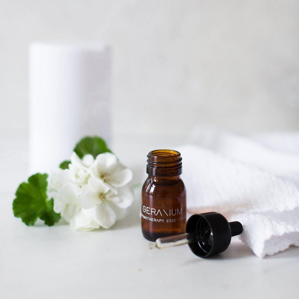 Een RainPharma Essential Oil Geranium amberkleurig druppelflesje staat naast een witte handdoek, delicate witte bloemen en een groen blad - ideaal voor gebruik in uw aromadiffuser om te genieten van de verfijnde geur van geranium.