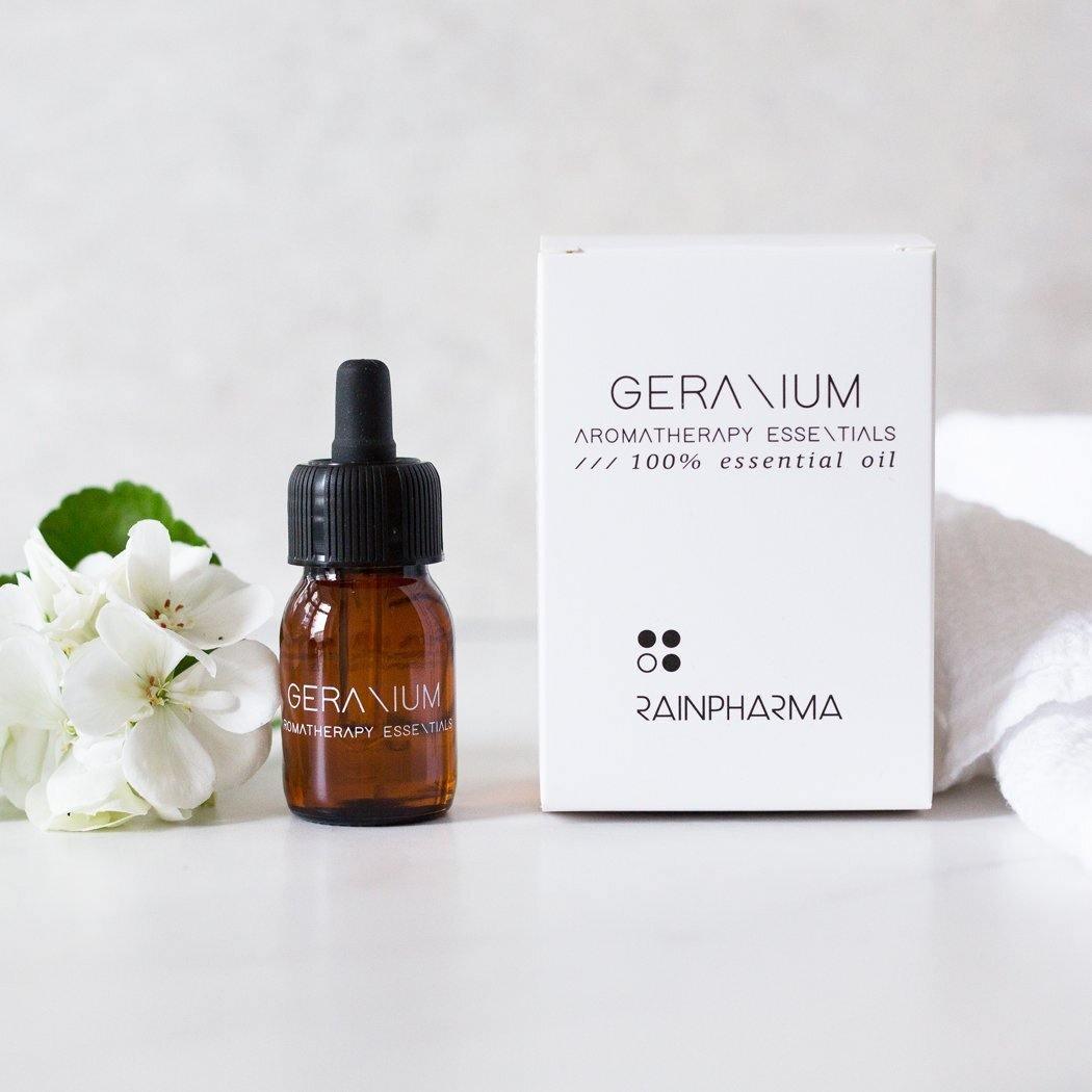Een bruin RainPharma "Essential Oil Geranium" druppelflesje zit naast een wit doosje, een delicate witte bloem en een handdoek - ideaal om deze geranium essentiële olie toe te voegen aan je aromadiffuser.