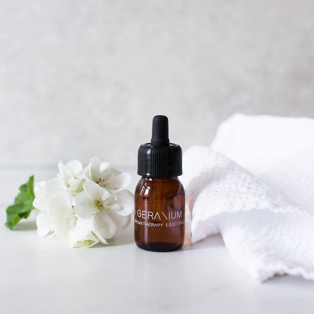 Een klein bruin glazen druppelflesje met het label "Essential Oil Geranium" van RainPharma staat naast witte bloemen en een gevouwen witte handdoek, perfect om te gebruiken in je aroma diffuser met een kalmerende geranium geur.