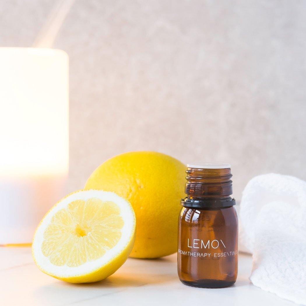 Een flesje RainPharma etherische olie citroen staat sierlijk naast een hele en een half citroenschijfje op een licht oppervlak, badend in zacht, gloeiend licht, waardoor de essentie van aromatherapie wordt vastgelegd.