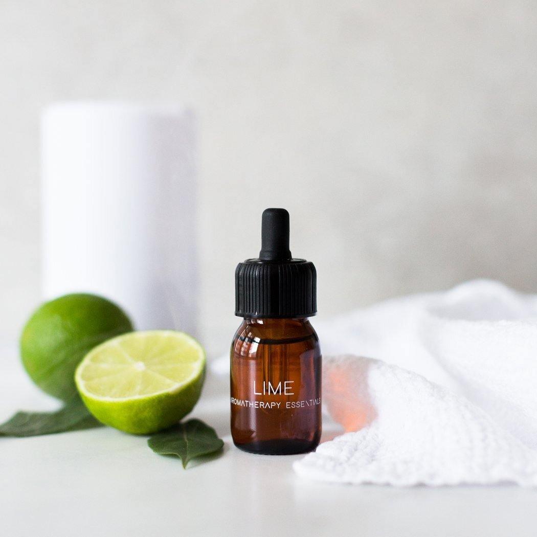 Een bruin RainPharma "Essential Oil - Lime 30 ml" flesje zit naast een gehalveerde limoen, hele limoen, groene blaadjes, witte handdoek en cilinder - perfect voor gebruik in je aroma diffuser of als verfrissende limoen geur essentiële olie.