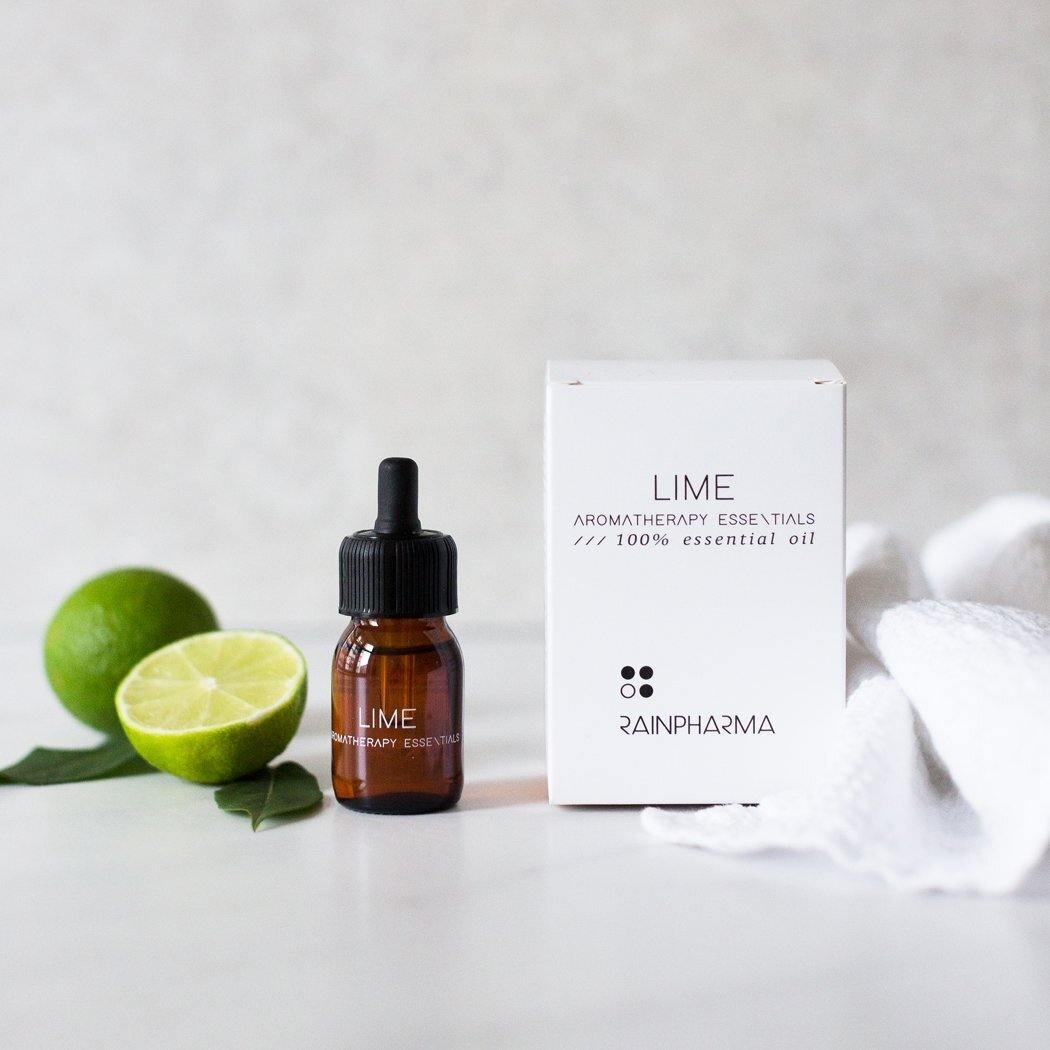 Een flesje etherische olie limoen van RainPharma met een druppelaar staat naast een doosje met het label "Lime Aromatherapy Essentials" en een gehalveerde limoen op een wit oppervlak. Ideaal voor gebruik in een diffuser om optimaal gebruik te maken van de therapeutische eigenschappen.