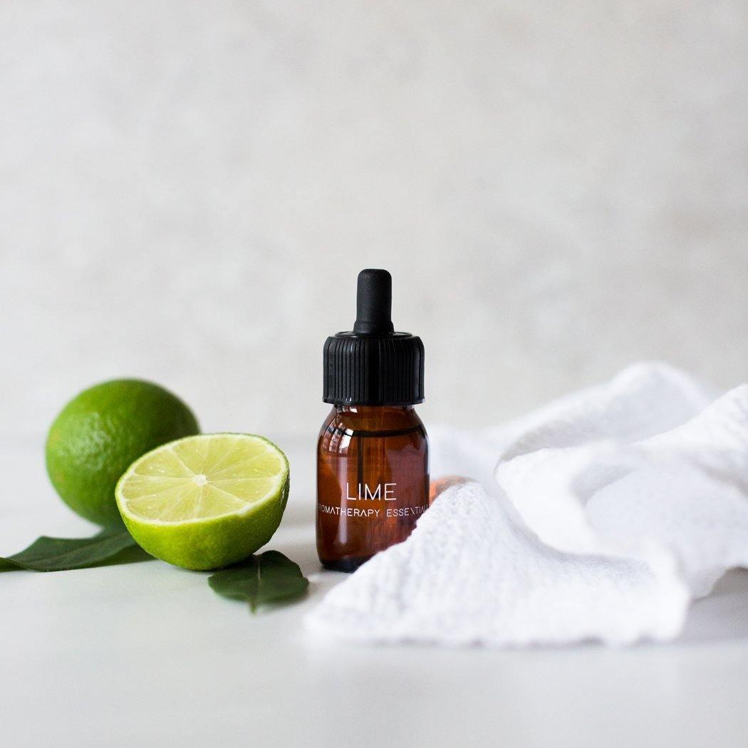 Een bruine fles met het opschrift "Essential Oil Lime" van RainPharma staat naast een gehalveerde limoen, een hele limoen en een witte doek op een licht oppervlak, perfect om uw aroma diffuser van zijn therapeutische eigenschappen te laten genieten.