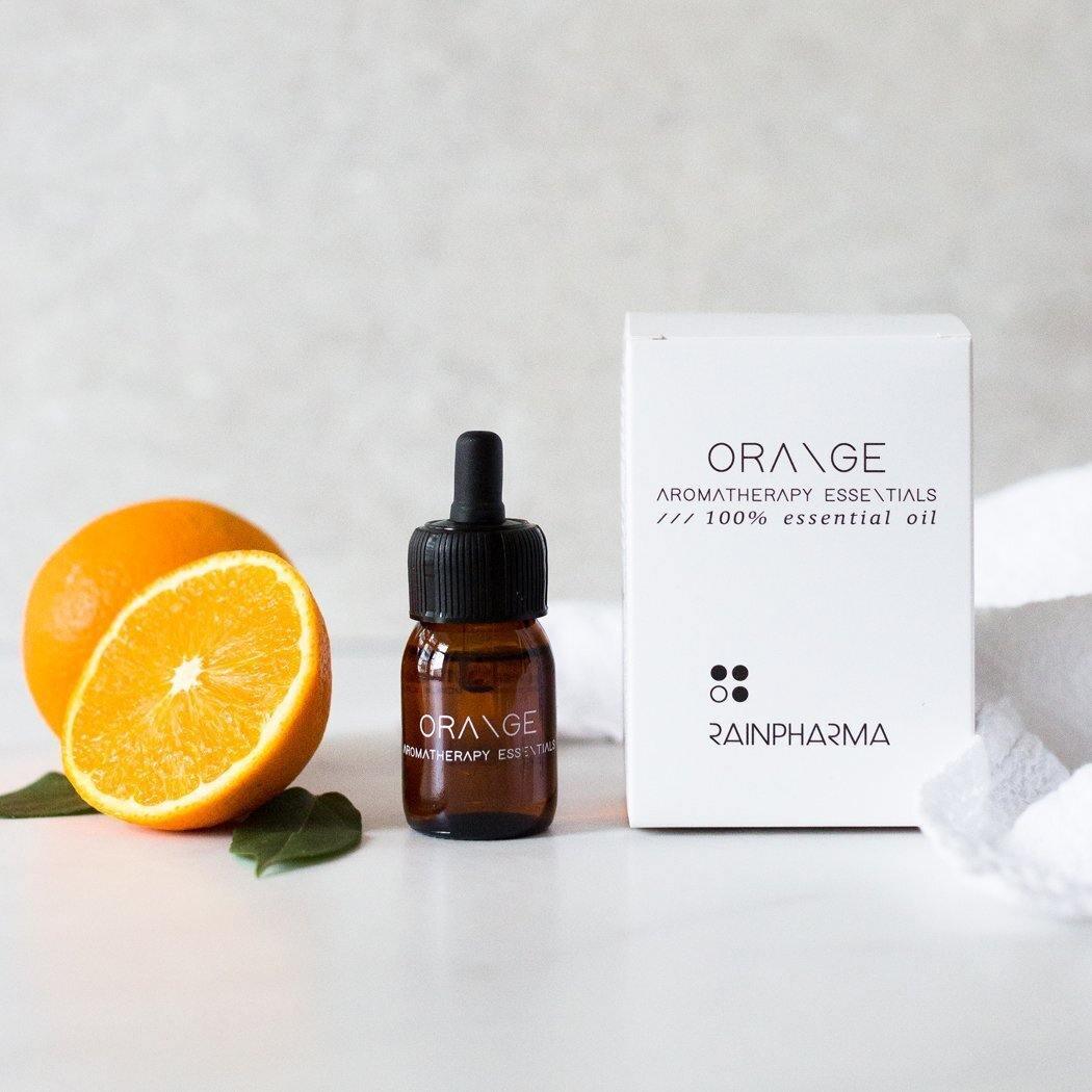 Een flesje etherische olie van RainPharma, sinaasappel, staat naast een sinaasappel, een doorgesneden helft en een wit doosje met het opschrift &