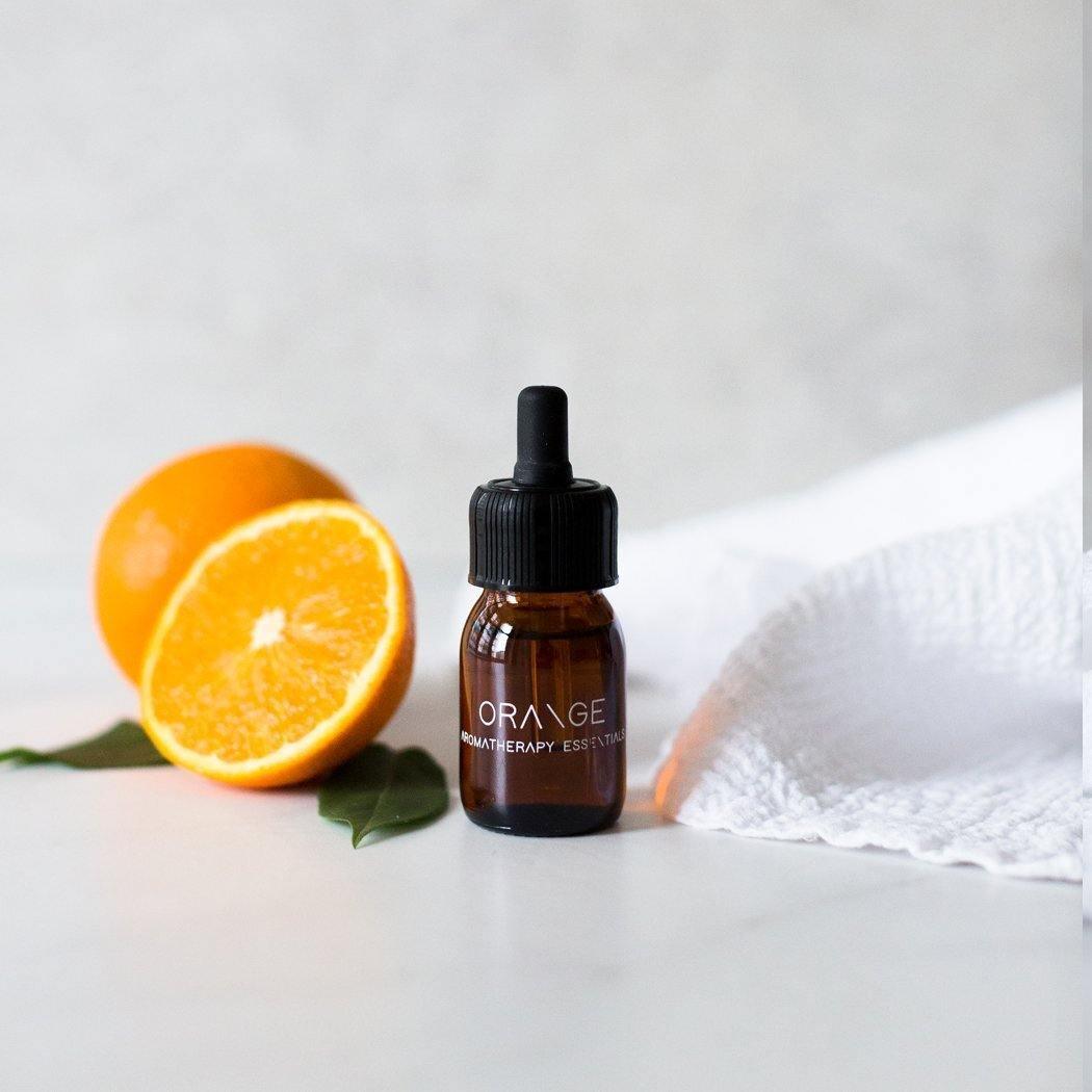 Een klein bruin flesje met het label "Essential Oil Orange" van RainPharma staat naast een sinaasappel en een gehalveerde sinaasappel op een wit oppervlak, en straalt een levendige kleur uit. De premium kwaliteit versterkt de therapeutische ambiance tegen de serene witte stoffen achtergrond.