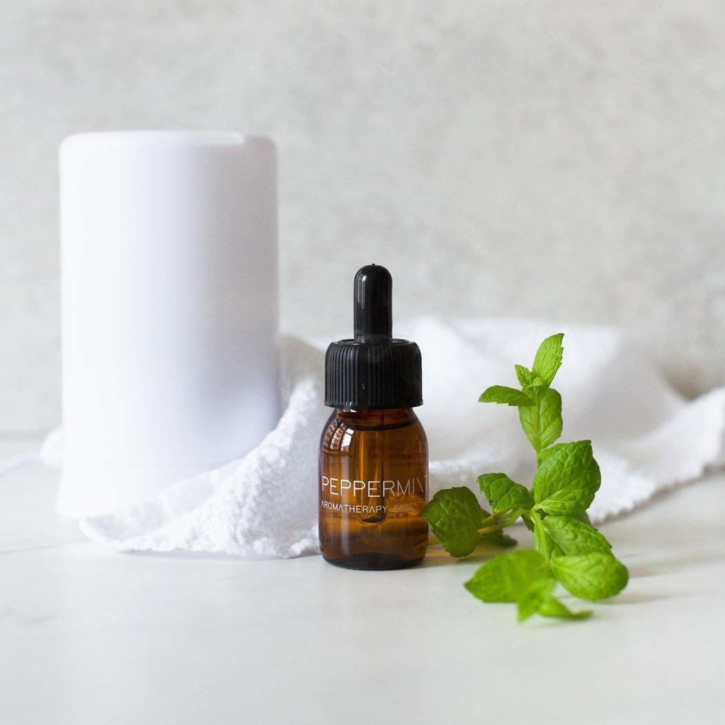 Een bruin druppelflesje met het label "RainPharma Essential Oil Peppermint" staat op een oppervlak met verse muntblaadjes, een witte handdoek en een cilindrische diffuser. Deze essentiële olie biedt therapeutische eigenschappen die uw aroma diffuser ervaring verbeteren.