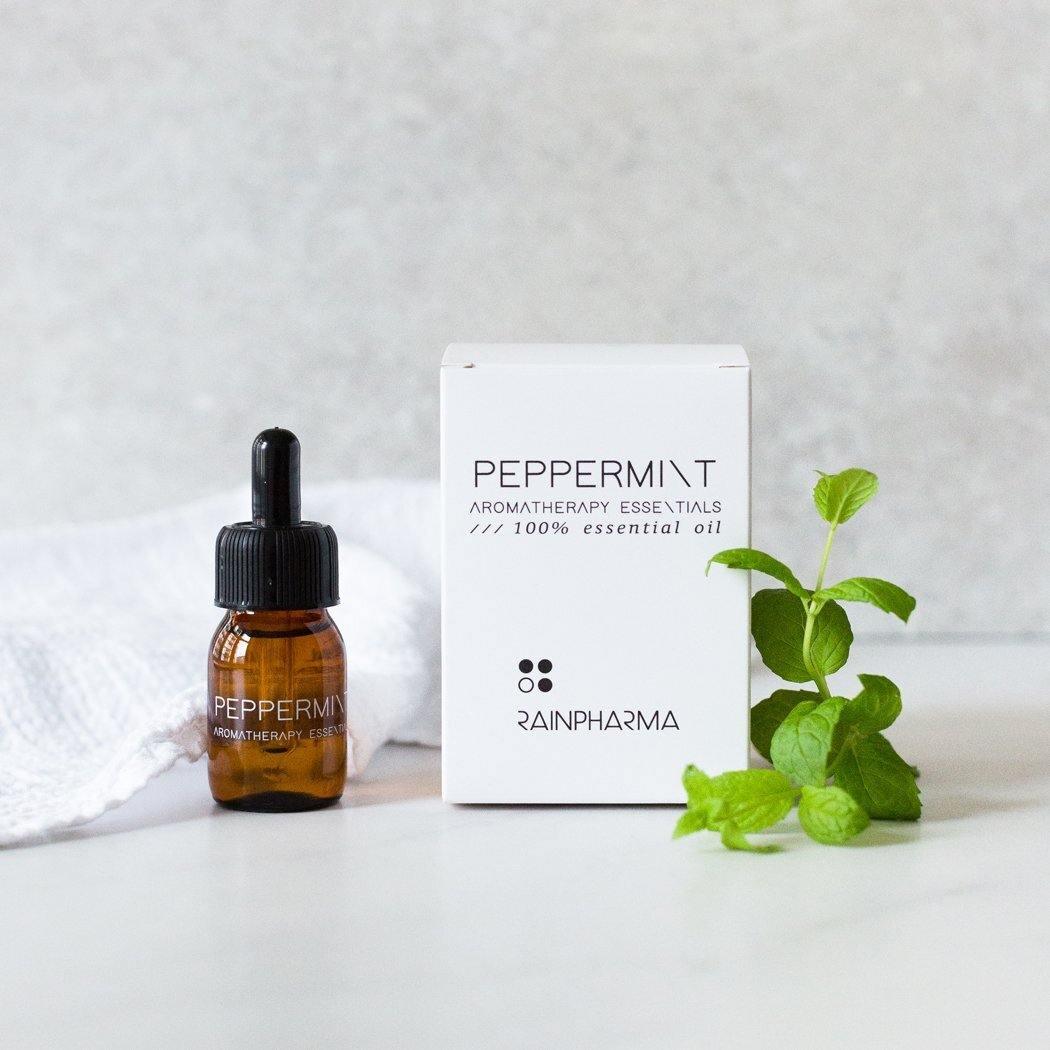 Een klein bruin flesje met een druppelaar bevat "RainPharma Essential Oil Peppermint" en staat naast een witte doos met het label "Peppermint Aromatherapy Essentials", vergezeld door een takje verse munt. Perfect voor aroma diffusers, deze olie biedt indrukwekkende therapeutische eigenschappen.