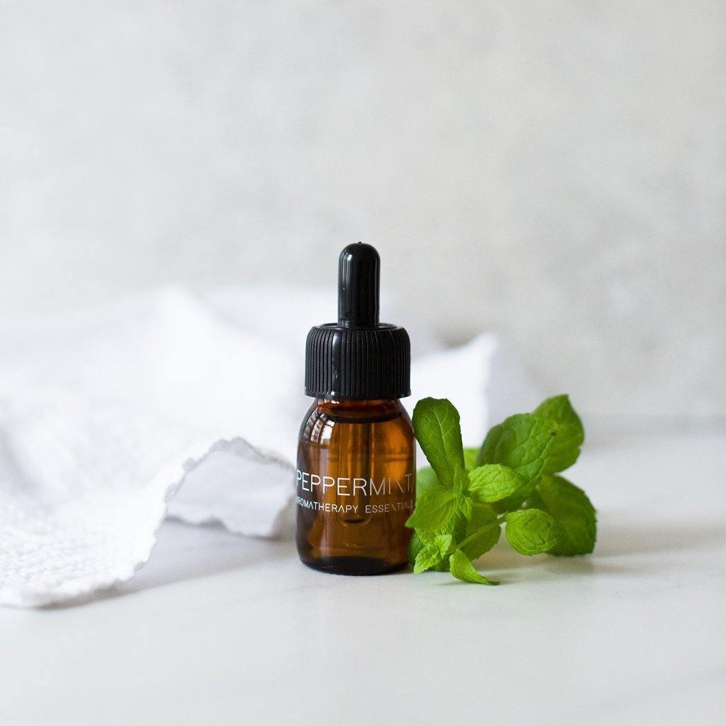 Een klein amberkleurig flesje van RainPharma, gelabeld "Essential Oil Peppermint" met een druppelaar, staat naast verse munt op een witte doek. Ideaal voor uw Aroma Diffuser, het benadrukt therapeutische voordelen.