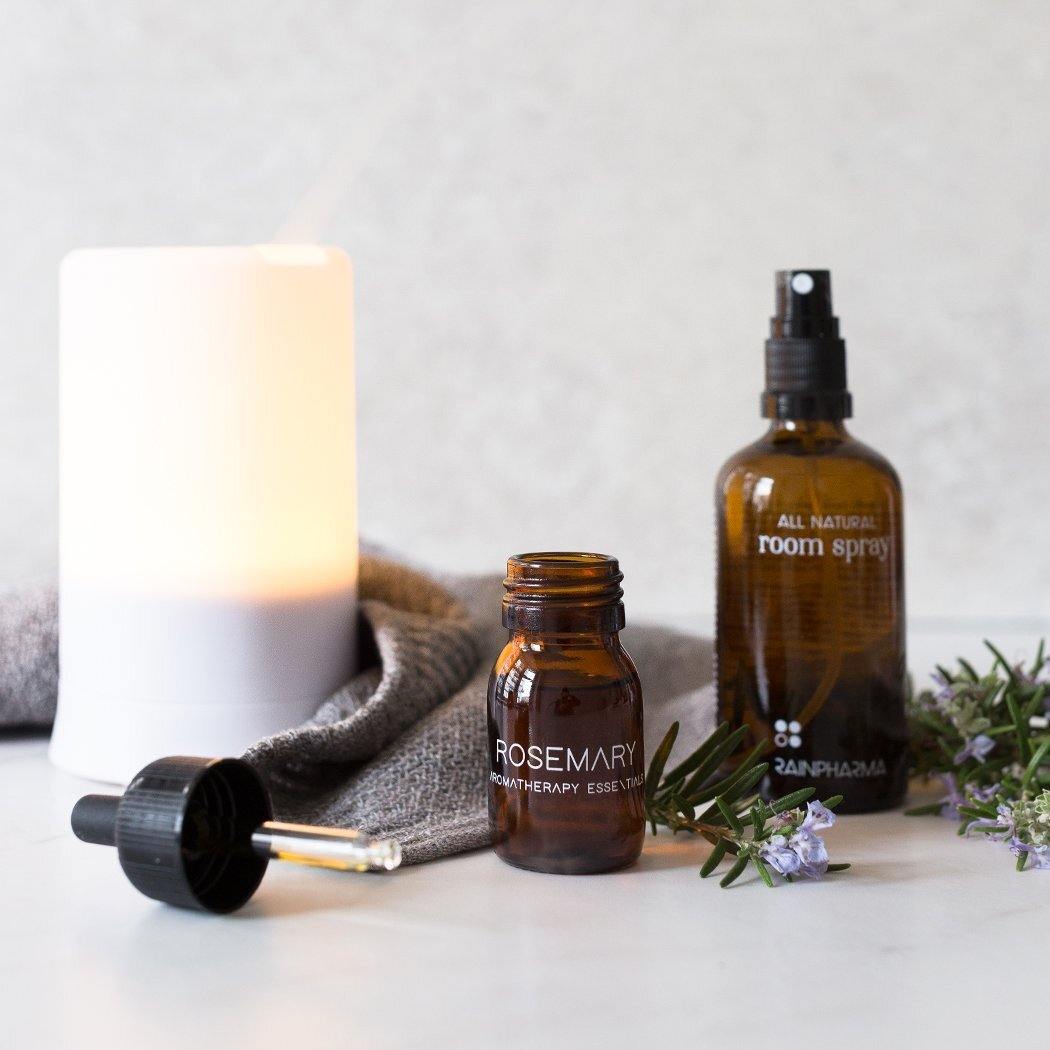 Een druppelaar voor essentiële olie en rozemarijn staan naast een diffuser, een klein flesje met het label "RainPharma Essential Oil Rosemary" en een bruine verstuiver met het label "All Natural Room Spray", ideaal om thuis te genieten van de kalmerende effecten van aromatherapie.