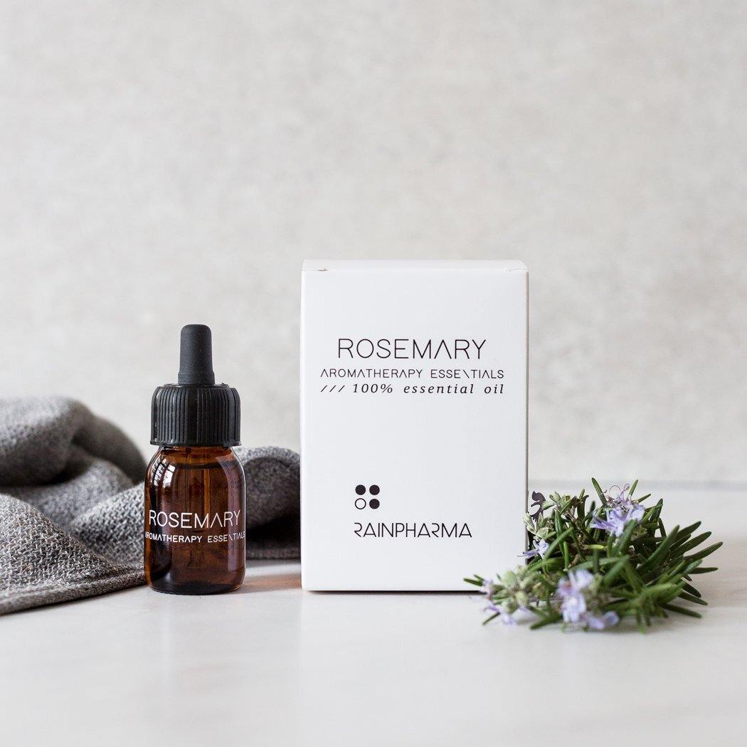 Een flesje etherische olie rozemarijn van RainPharma staat naast een doos met "Aromatherapy Essentials", samen met rozemarijn, op een wit oppervlak met grijze stoffen accenten.