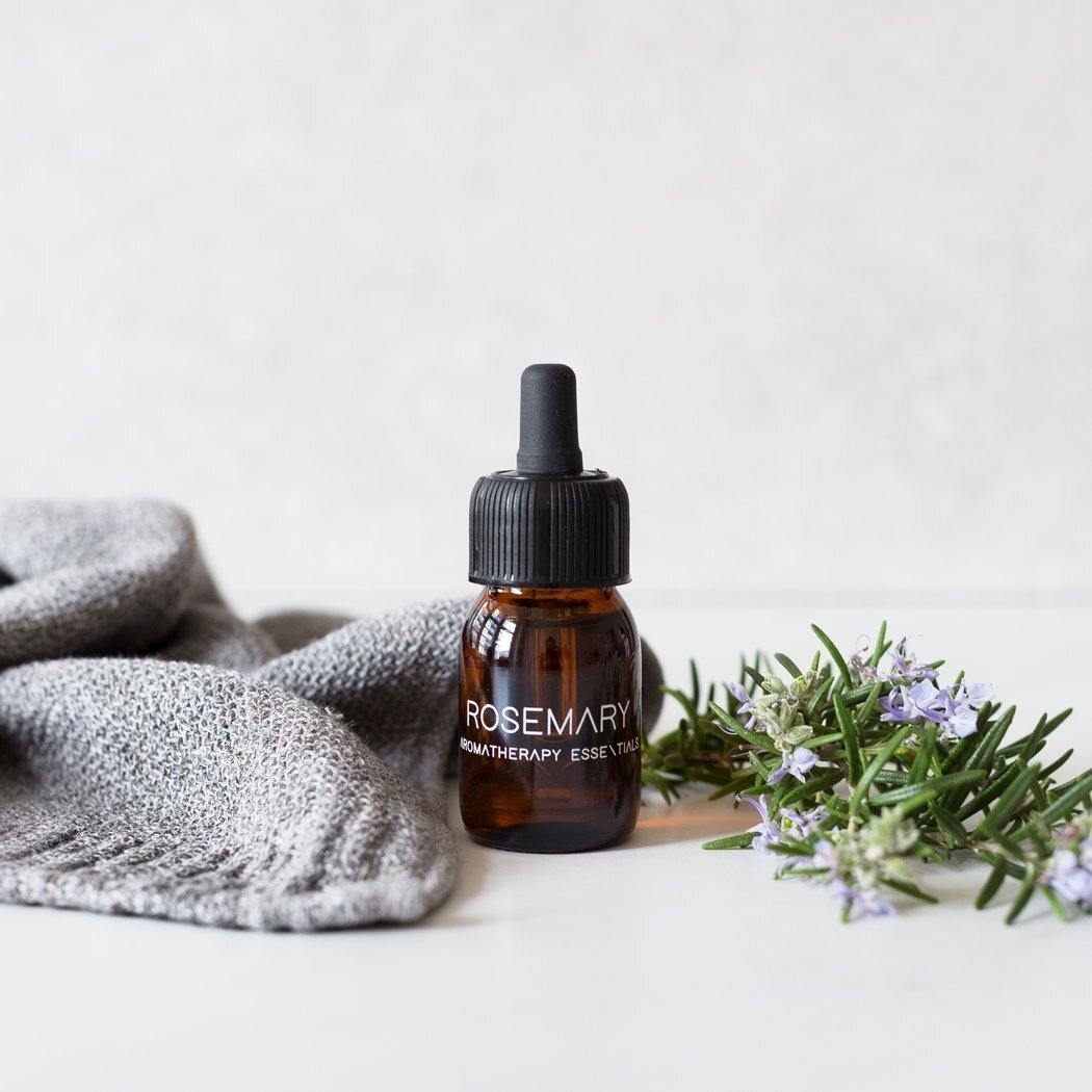 Een bruin flesje met het opschrift "Essentiële olie Rozemarijn" van RainPharma heeft een druppeldop, een takje rozemarijn en een grijze doek op een licht oppervlak.