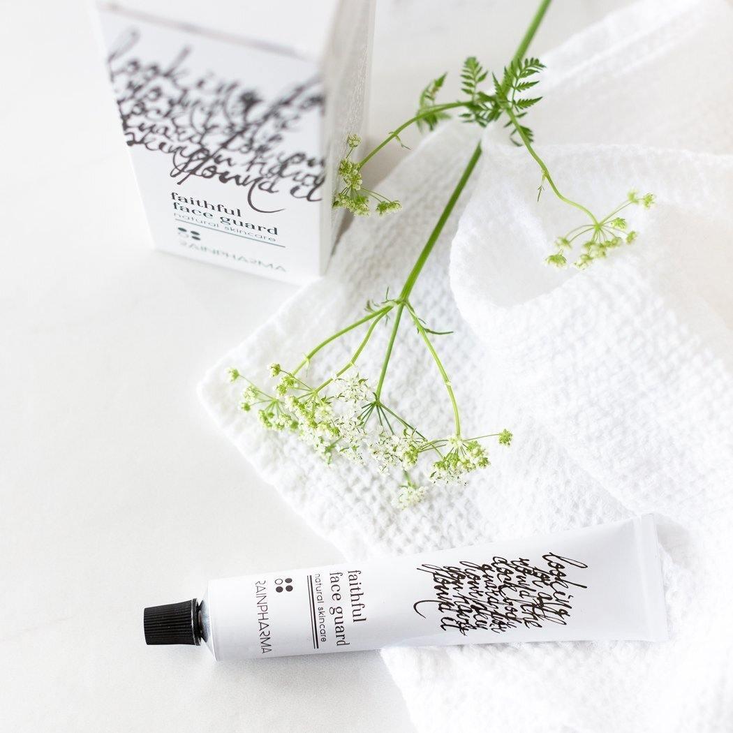 Een tube met het label "Faithful Face Guard (dag- en nachtcrème)" van RainPharma rust op een witte handdoek met groene bloemen, een verwijzing naar de formule verrijkt met Tamanu-olie. Een doos met cursieve tekst is elegant op de achtergrond geplaatst.