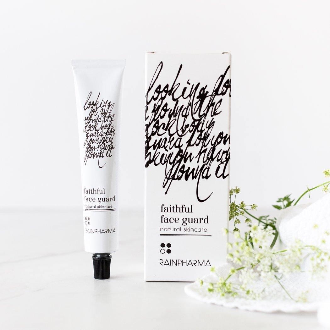 Een tube en doosje Faithful Face Guard (dag- en nachtcrème) huidverzorging van RainPharma liggen op een wit oppervlak, verrijkt met Tamanu-olie en rode algenextract, vergezeld door decoratieve bloemen.
