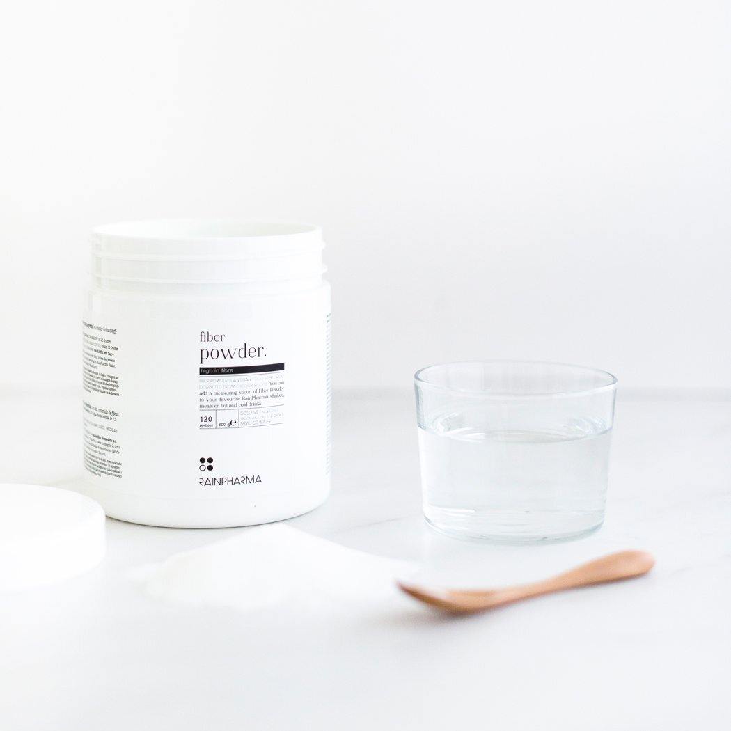 Een open verpakking RainPharma Fiber Powder staat naast een glas water, met een houten lepel lichtjes bedekt met het poeder op een witte achtergrond.